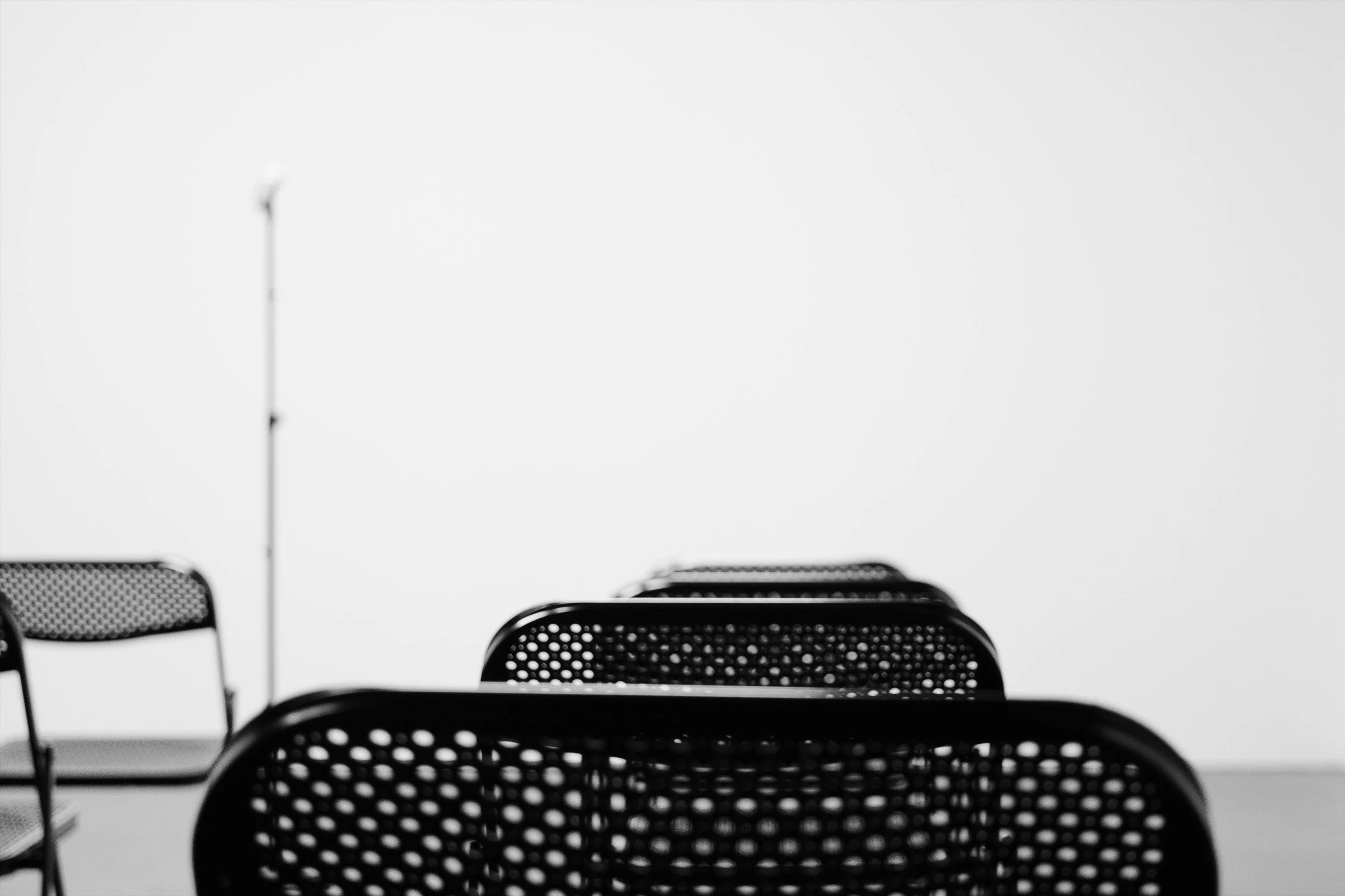 Empty black chairs with perforated seats and backs arranged in rows in front of a plain light-colored wall.