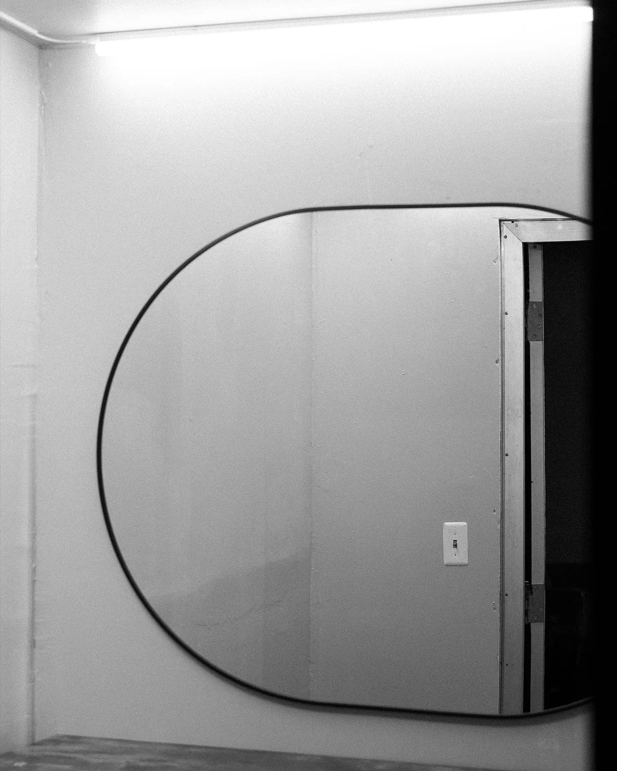 A mirror on a wall reflects a doorway with a metal frame, a light switch, and part of a ceiling with a long fluorescent light.