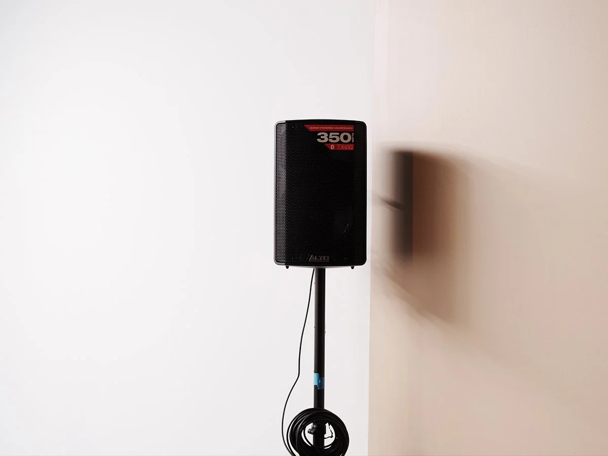 A black ALTO speaker on a black stand against a wall, with a wire hanging down and a coiled cable on the floor.