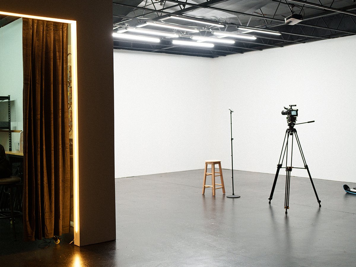 Empty video or photo studio with a stool, microphone, camera on tripod, and a dark ceiling with lighting. Partially visible room with a curtain and chairs.