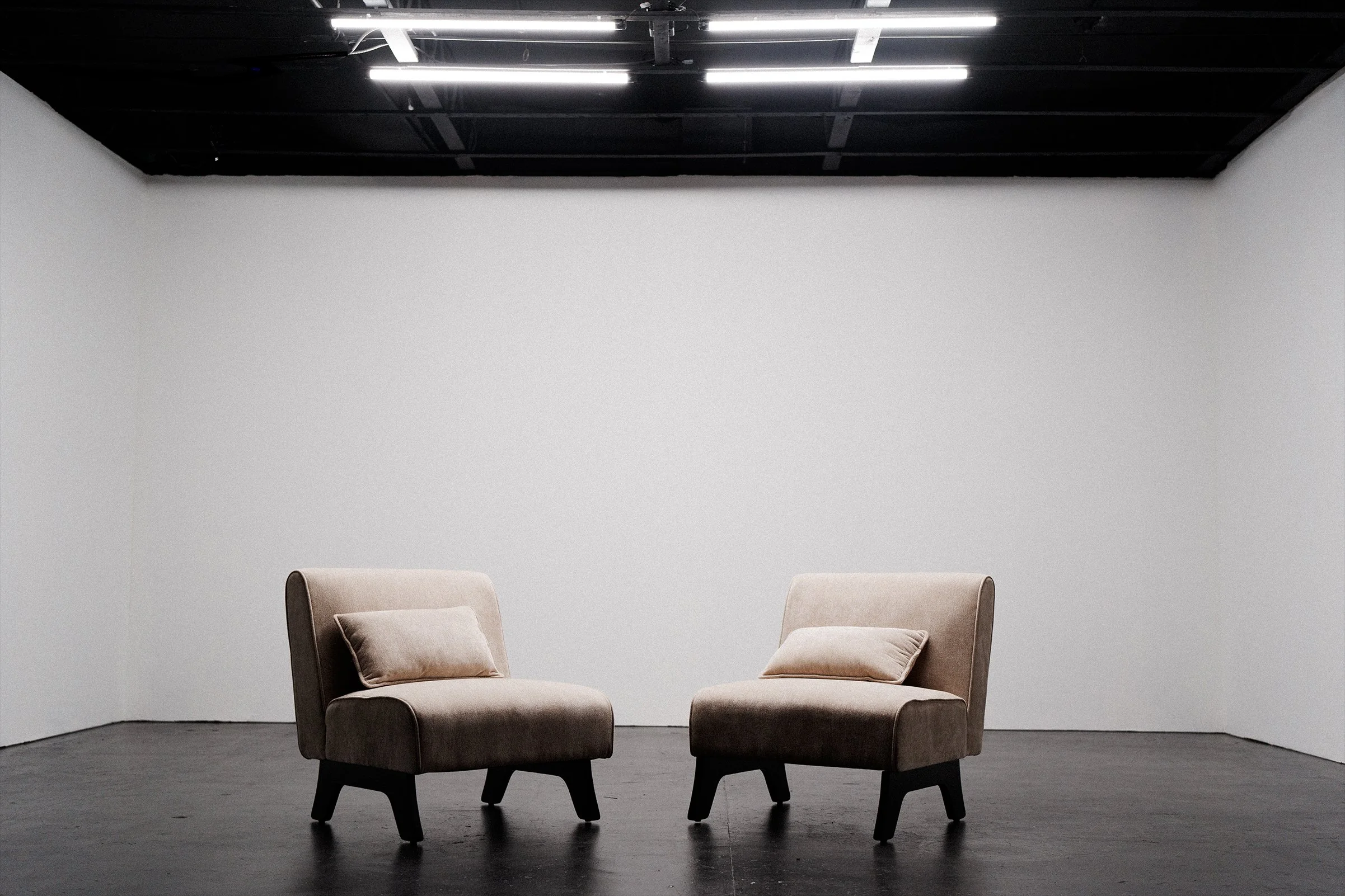Two beige armchairs with beige pillows in a minimalist room with gray walls and a black ceiling with modern lighting fixtures.