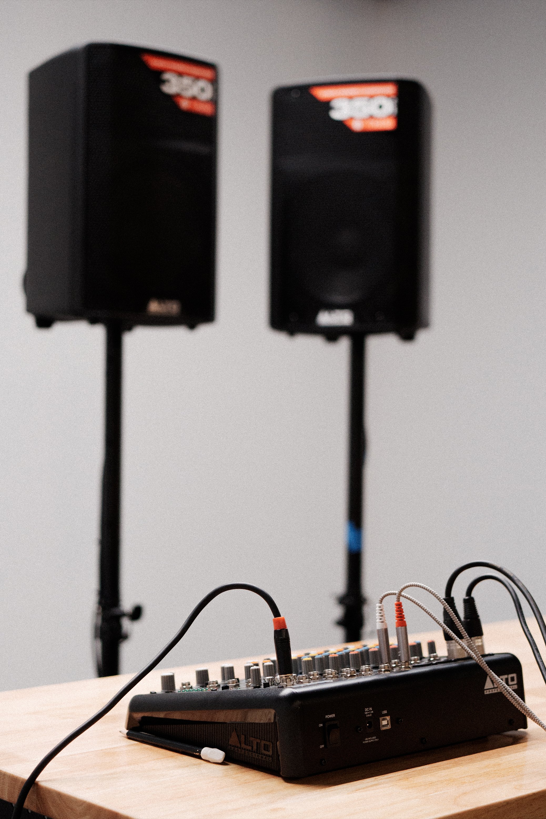 A music studio setup with two black speakers elevated on stands and a compact audio mixing console on a wooden table with various cables connected.