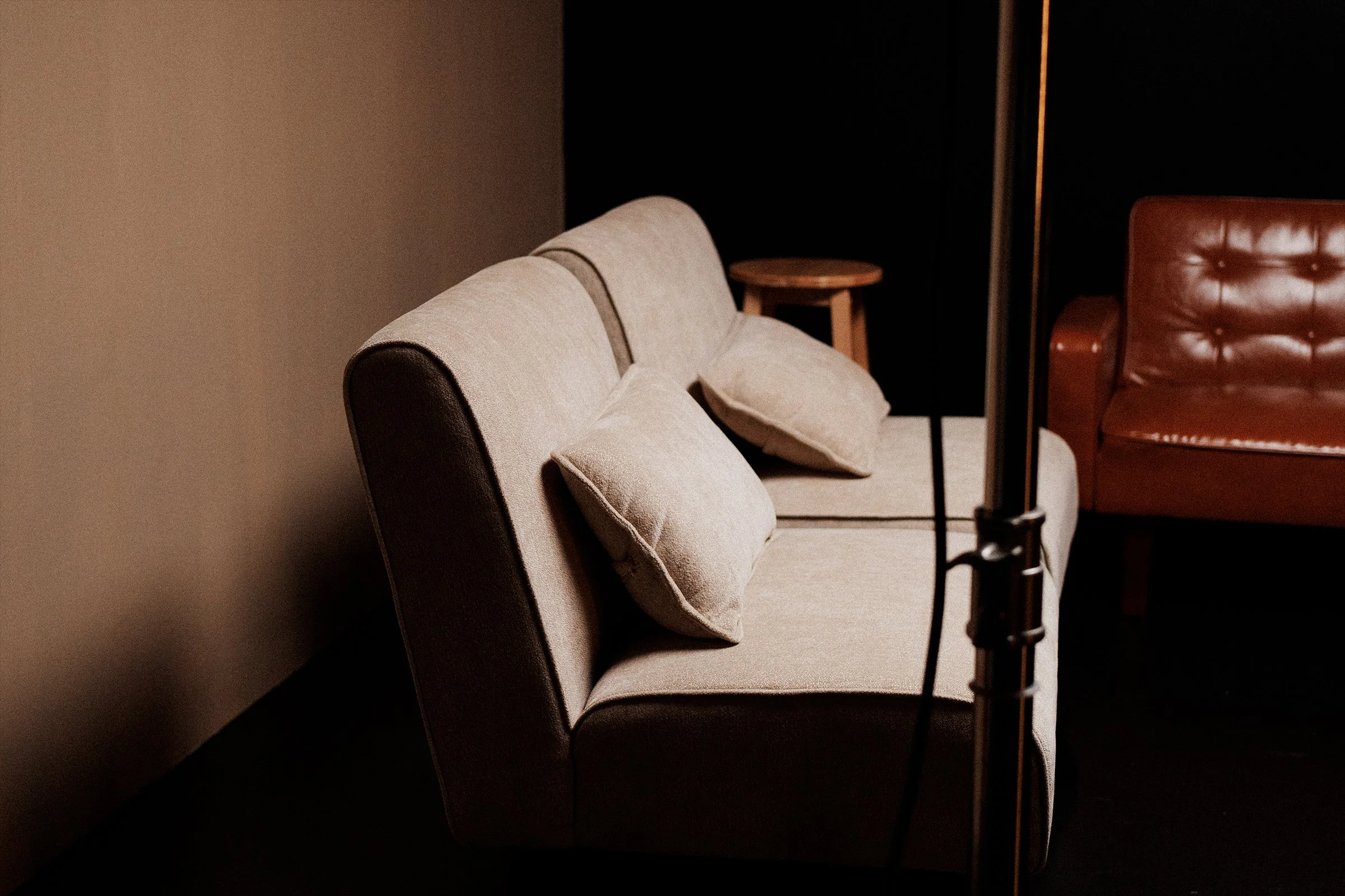 A beige sofa with three matching cushions next to a small wooden side table, with a tan leather armchair nearby in a dimly lit room.