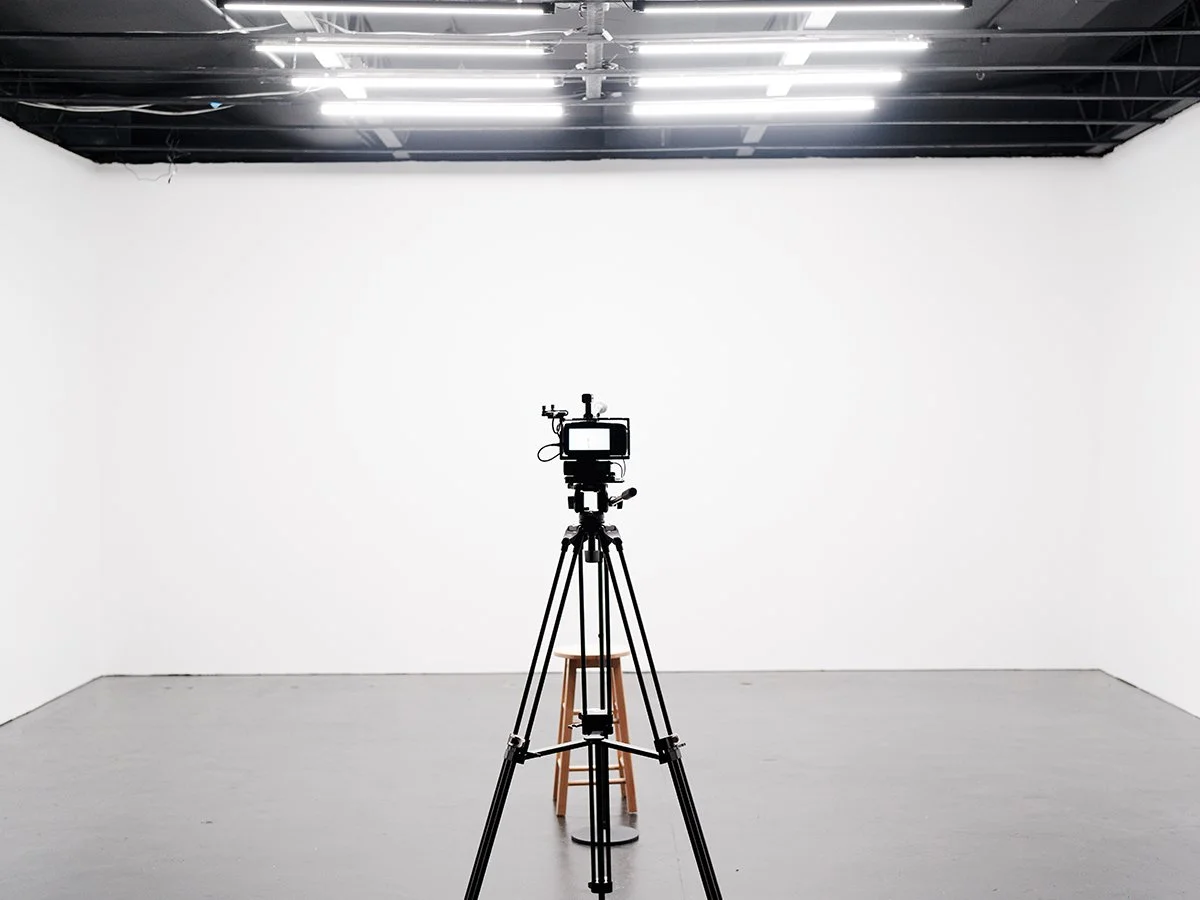 A camera on a tripod in a photography studio with white walls, gray floor, and studio lighting on the ceiling, and a wooden stool positioned in front of the camera.