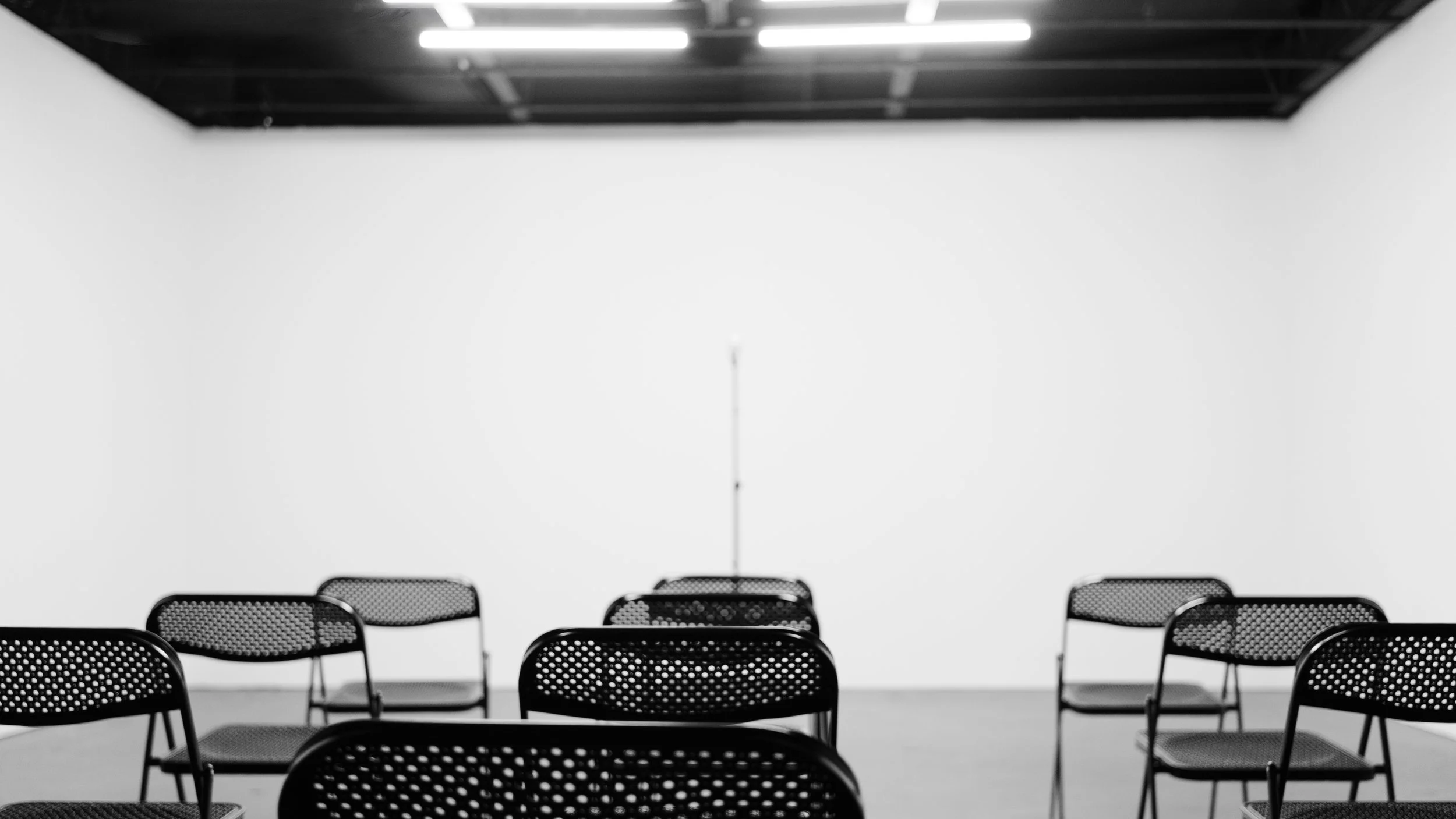 Empty room with rows of black metal chairs facing a blank white wall, ceiling with fluorescent lights, and a microphone stand in the center.