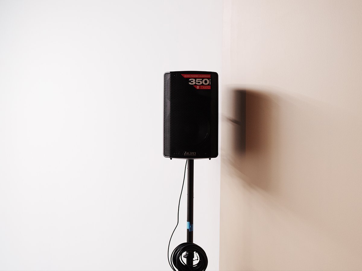 Black ALTO speaker mounted on a stand against a white and beige wall.