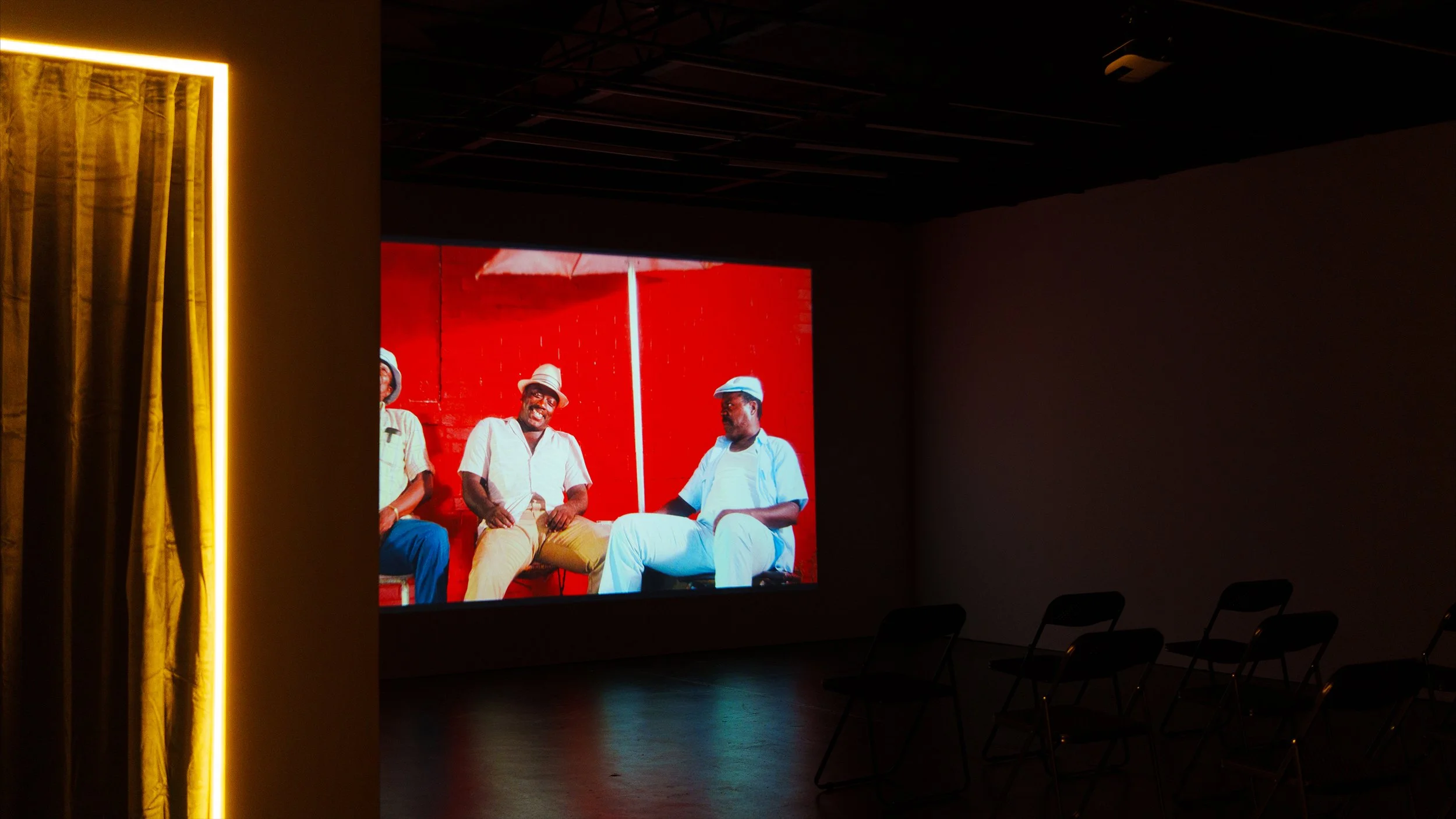 Large screen displaying a photo of four men sitting together, with three wearing white and one with a tan hat and glasses, against a red background in a dark room with chairs in the foreground.