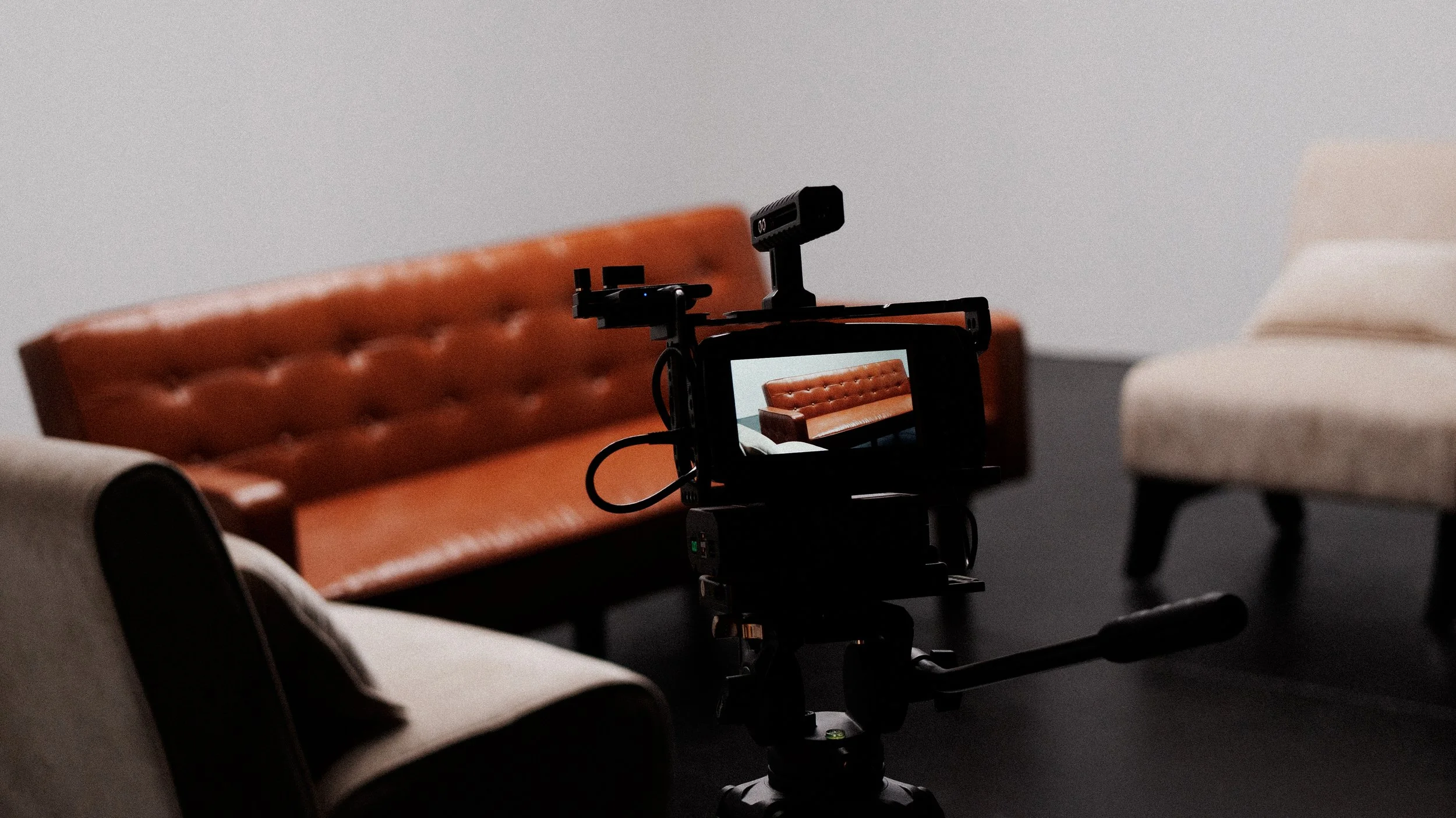 A camera on a tripod capturing a shot of a tan leather tufted sofa in a minimalist room with plain walls and a dark floor.