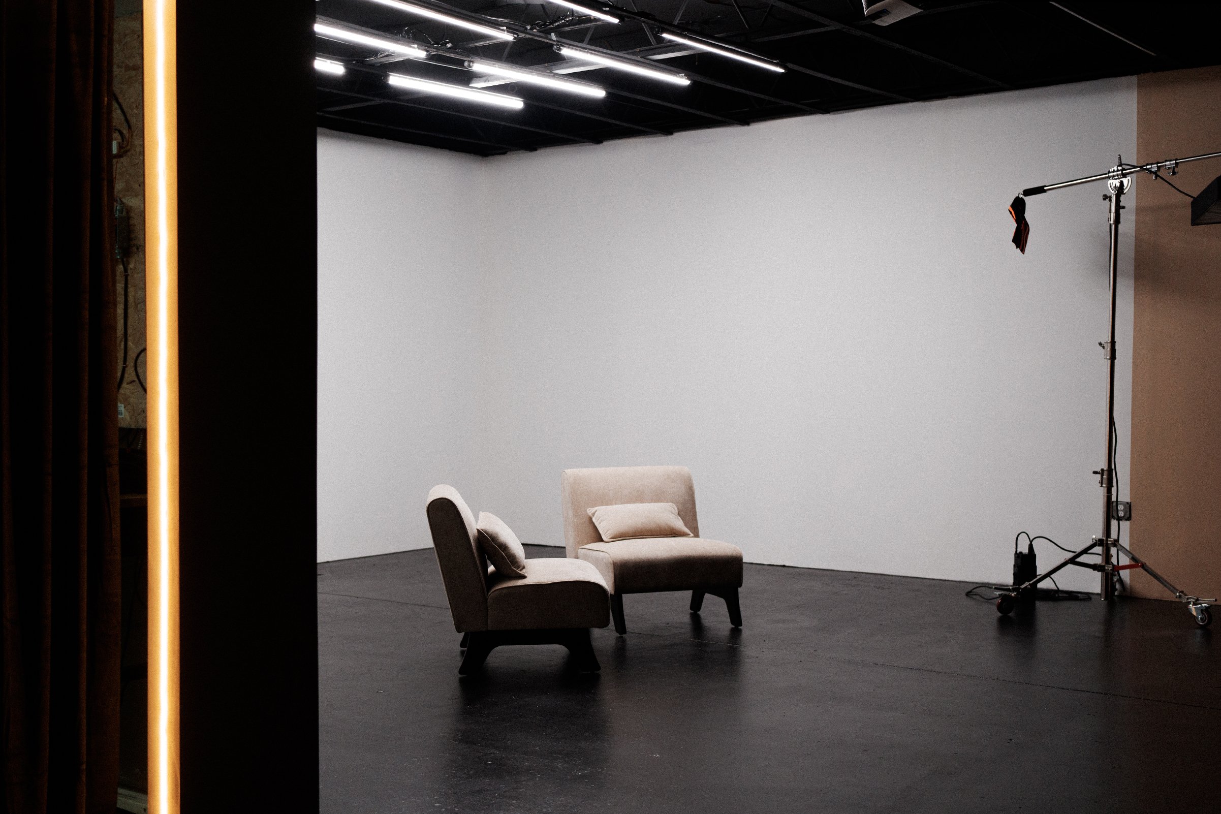 Photography studio setup with a white backdrop, two beige chairs with pillows, professional lighting equipment, and a dark floor.