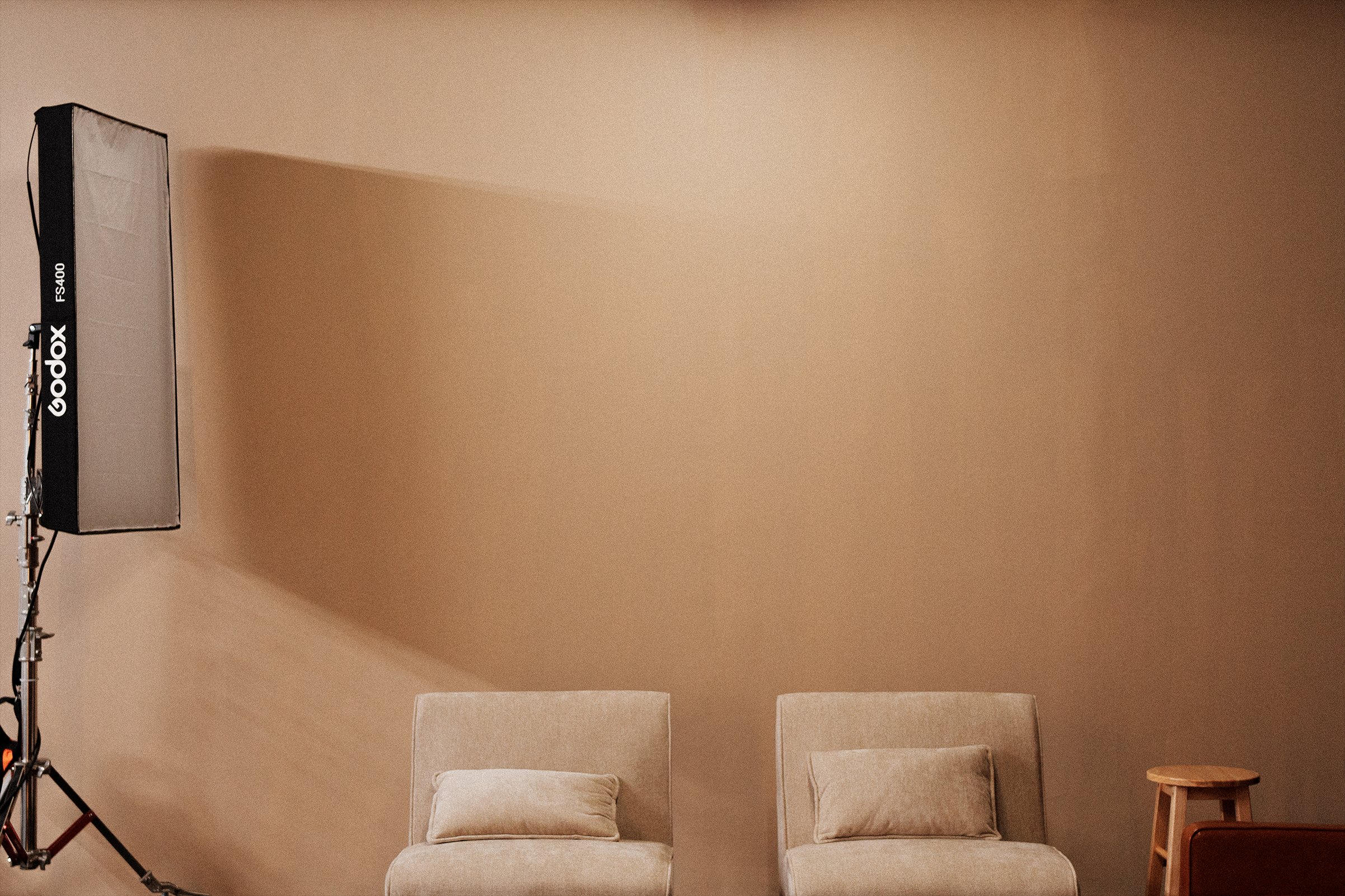 A beige room with two chairs and a small round wooden table. A soft pillow is on the left chair. There is a professional photography light on a stand on the left side of the image. The background wall is plain and light-colored.