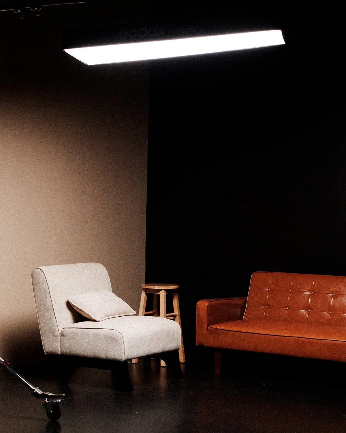 A living room set with a white armchair, a brown leather sofa, a small wooden stool, and a pillow on the armchair, in front of a black and beige wall under a bright overhead light.