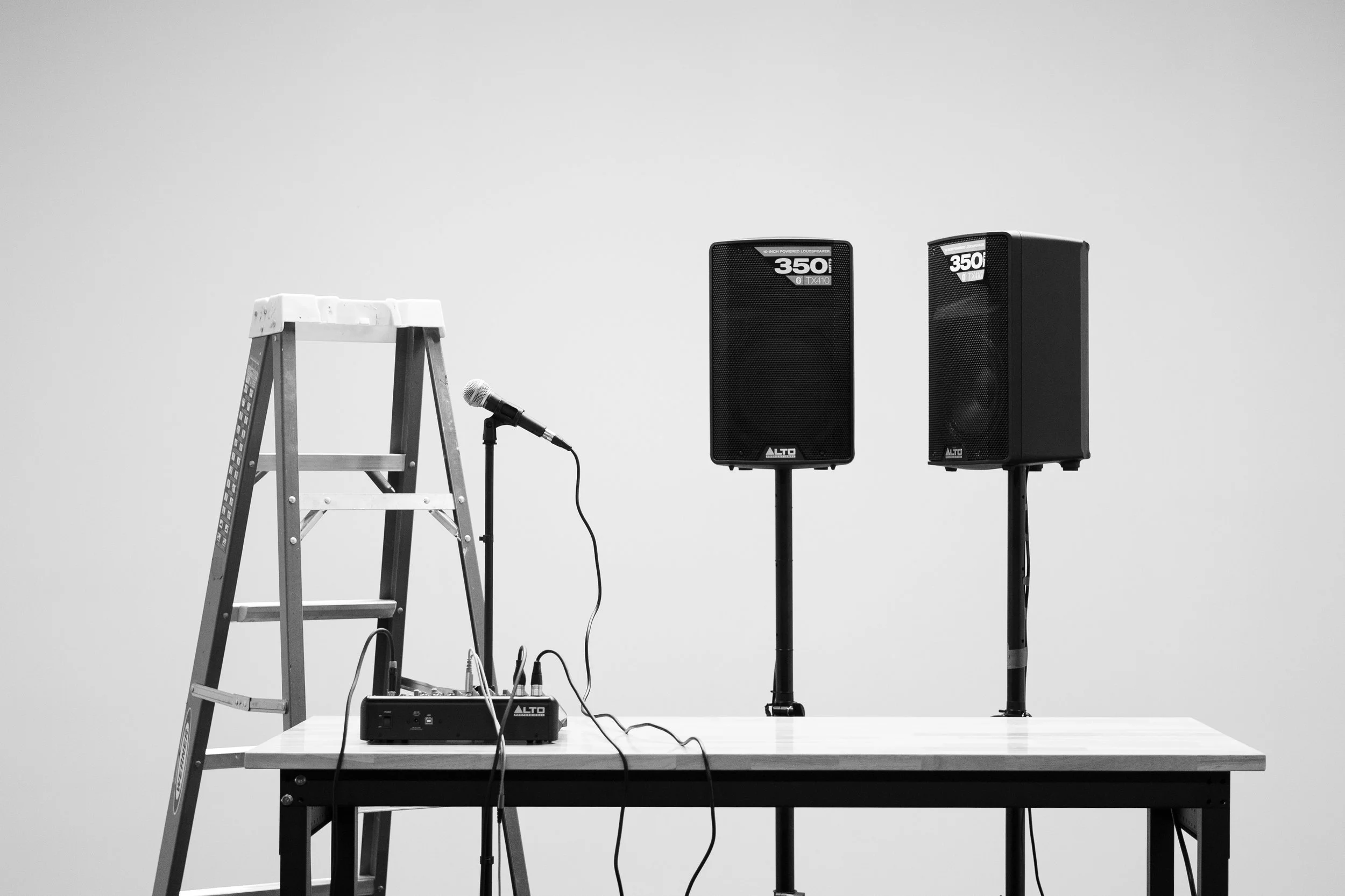 A sound setup with two large speakers on stands, a small audio mixer, a microphone on a stand, a wooden ladder, and a table. The background is plain and white.