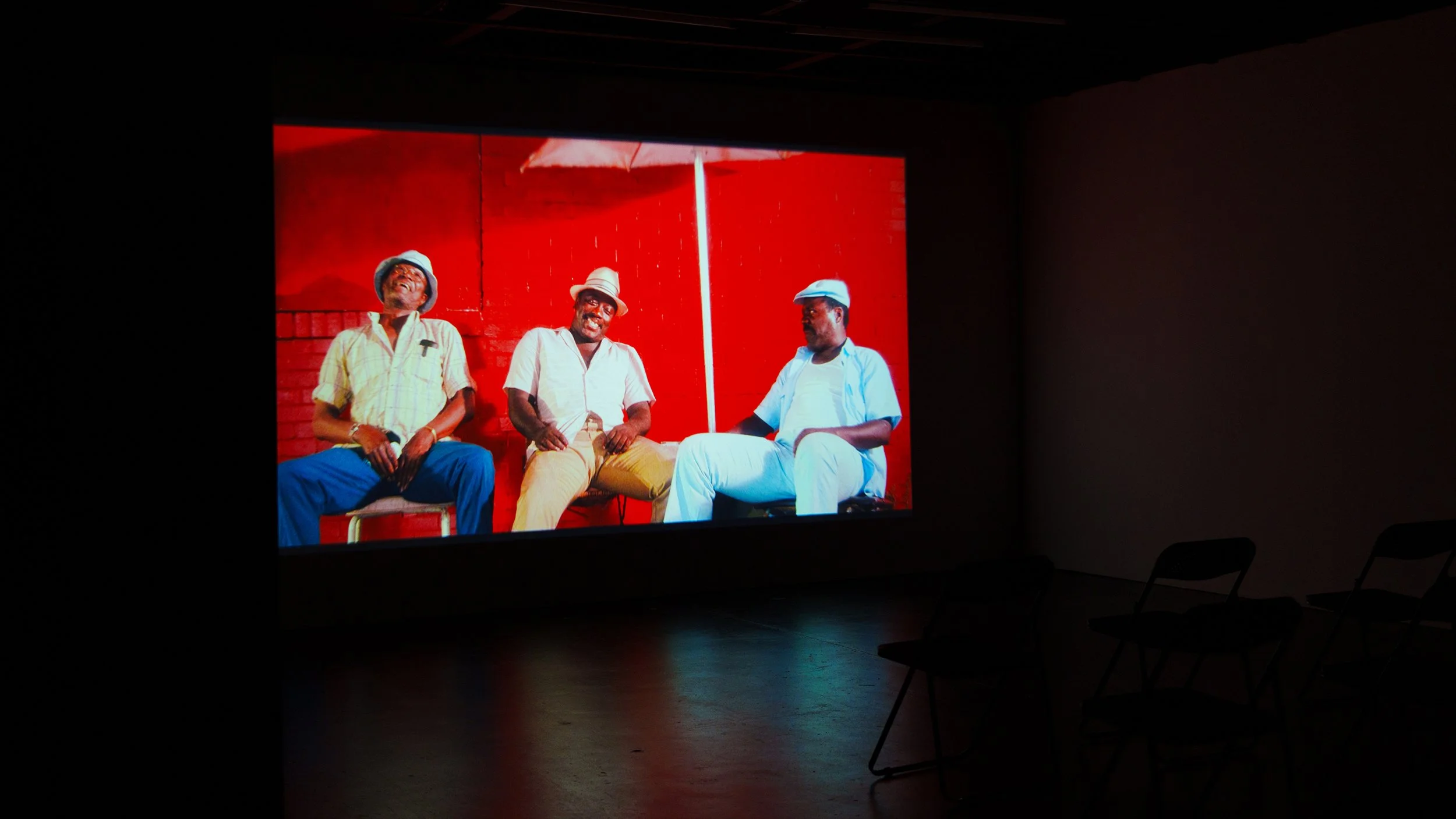 A large screen showing three men sitting and smiling against a red background with a red wall and a white parasol.