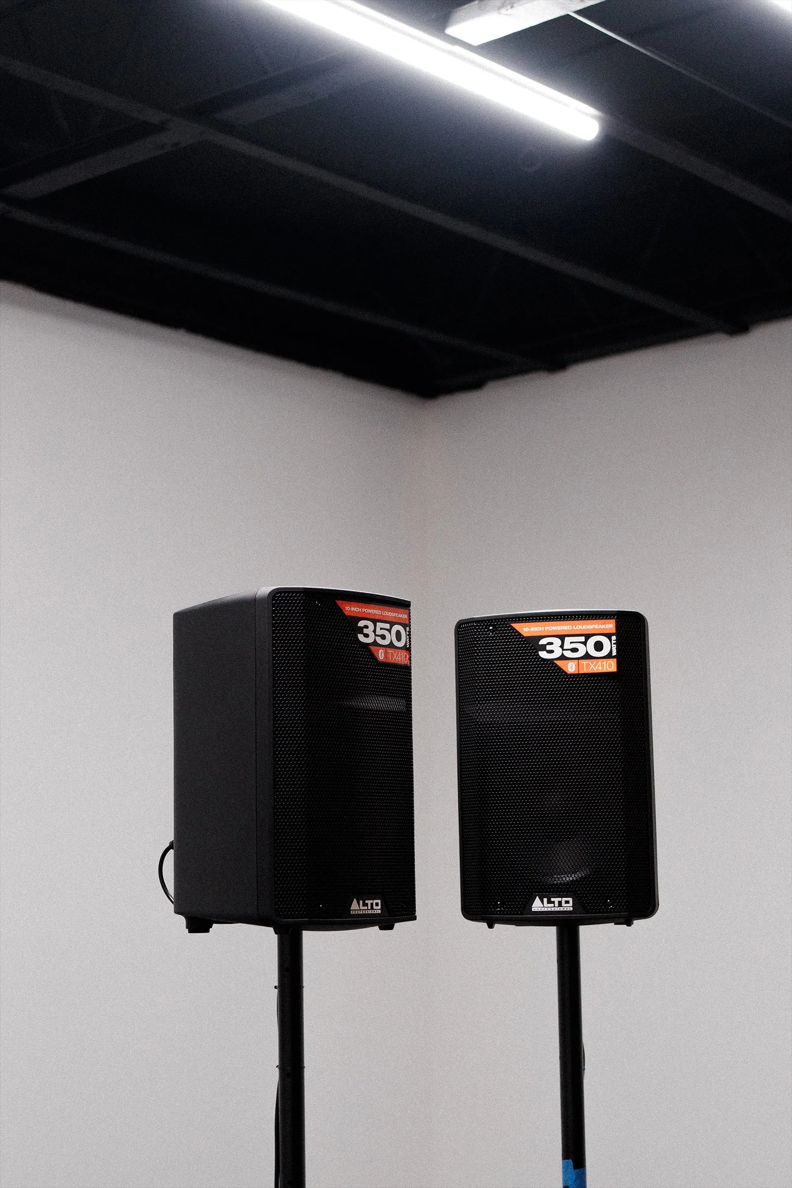 Two black ALTO speakers on stand, labeled '350' and 'TX410', in a room with grey walls and a white ceiling with fluorescent light.