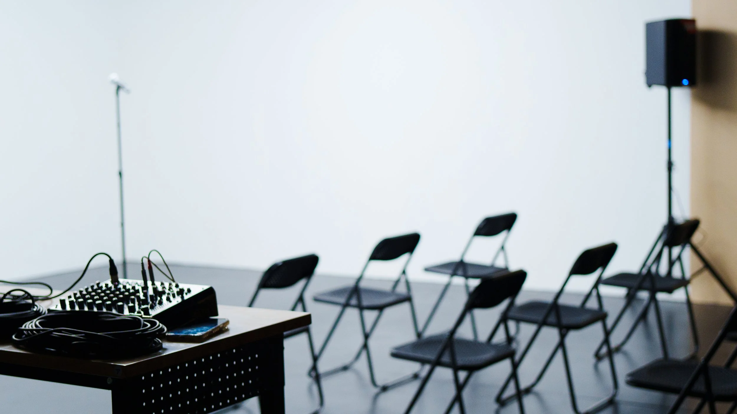 A room set up for a presentation or event with black chairs arranged in rows, a table with audio equipment including cables and a mixer, a microphone stand, and a large white wall or screen in the background.
