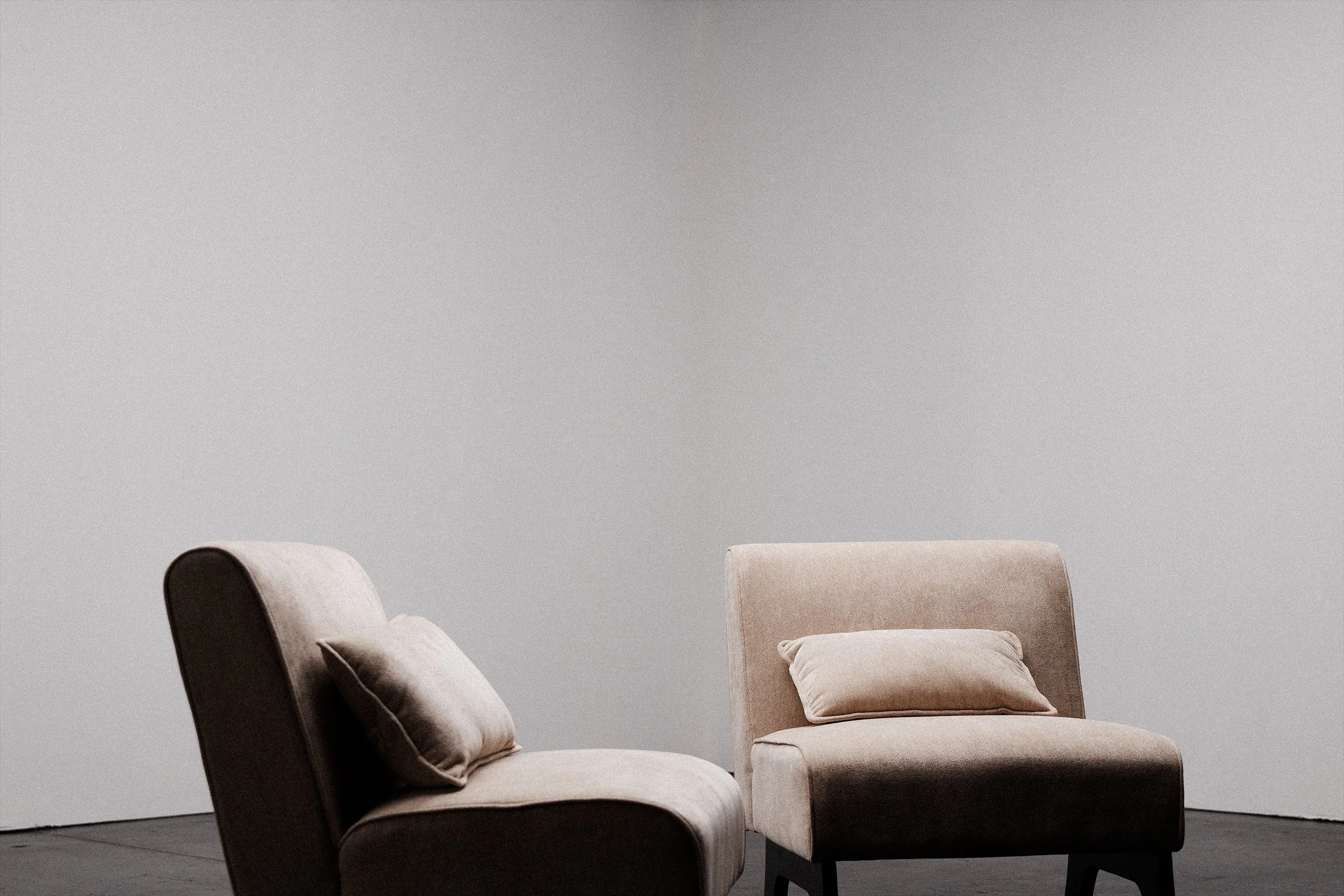 Two modern armchairs with pillows, one dark and one beige, positioned in front of a plain light gray wall.