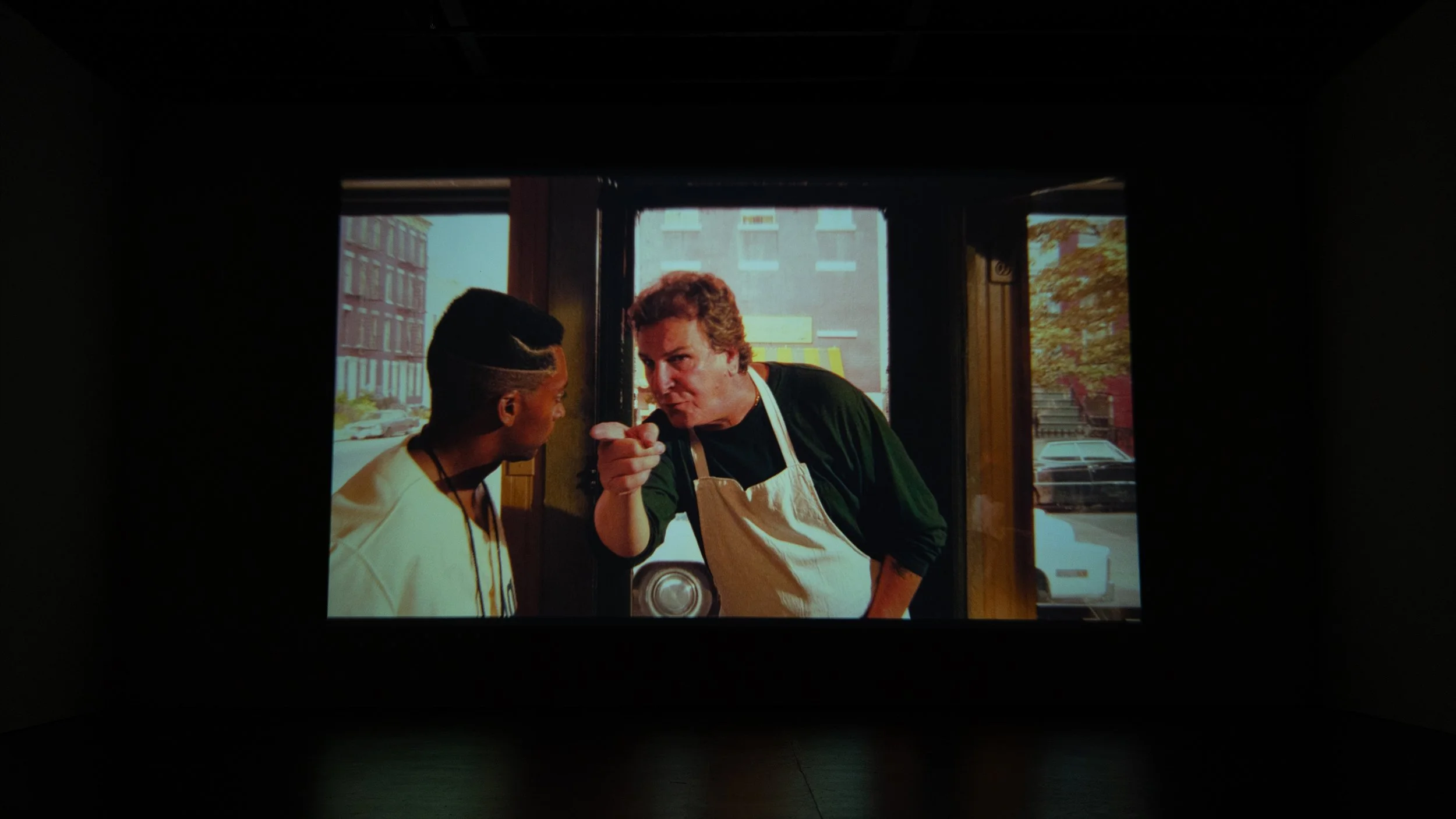 A scene from a movie or TV show with two men having an intense conversation. One man is wearing a white shirt, and the other man is wearing a dark jacket and an apron, pointing his finger at the other. They are standing by a window with a city street