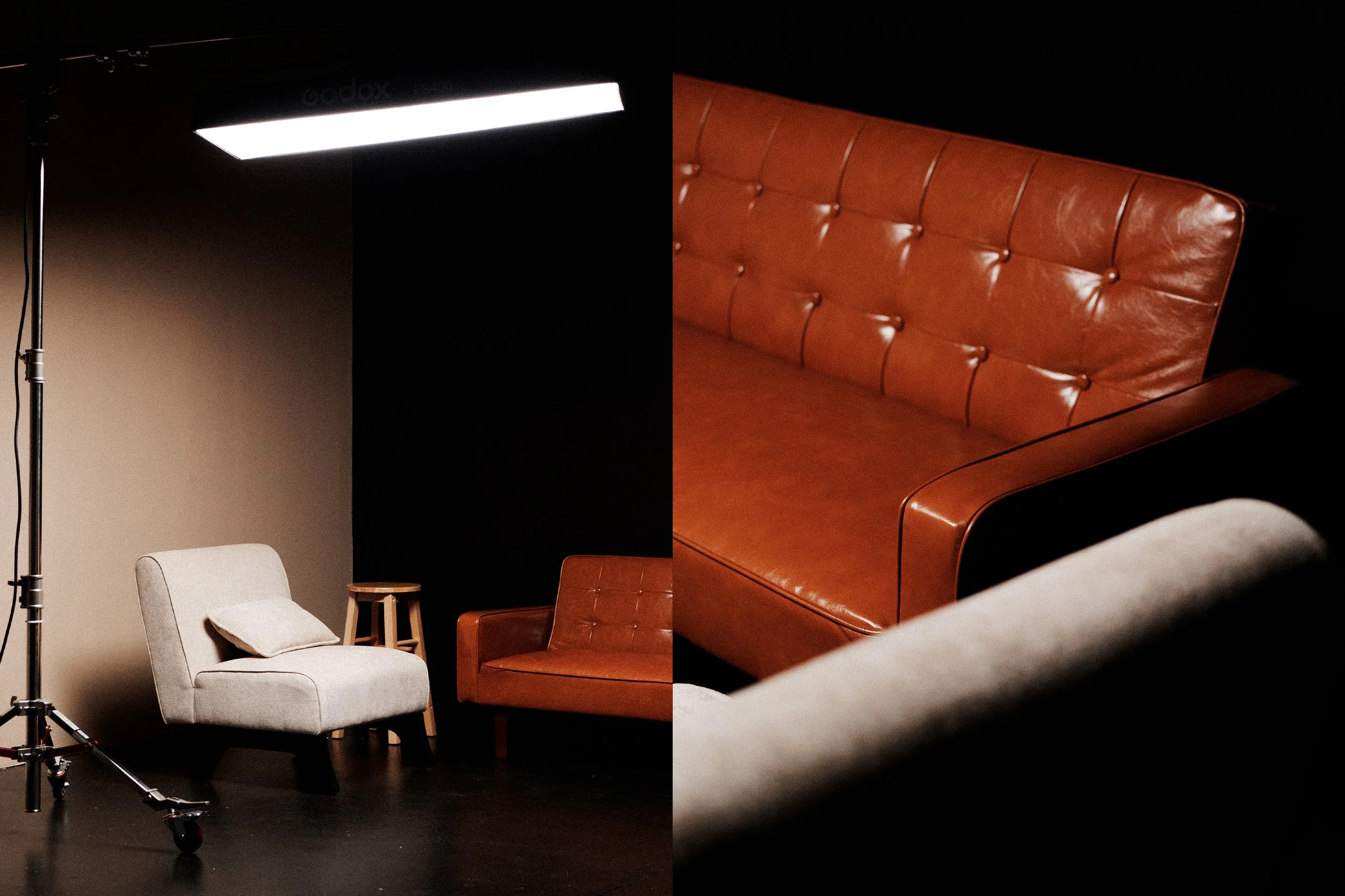 A photography studio setup with a professional lighting stand and a black backdrop, featuring various chairs including a white upholstered chair with a pillow, a brown leather chair, and a matching sofa positioned against the wall.
