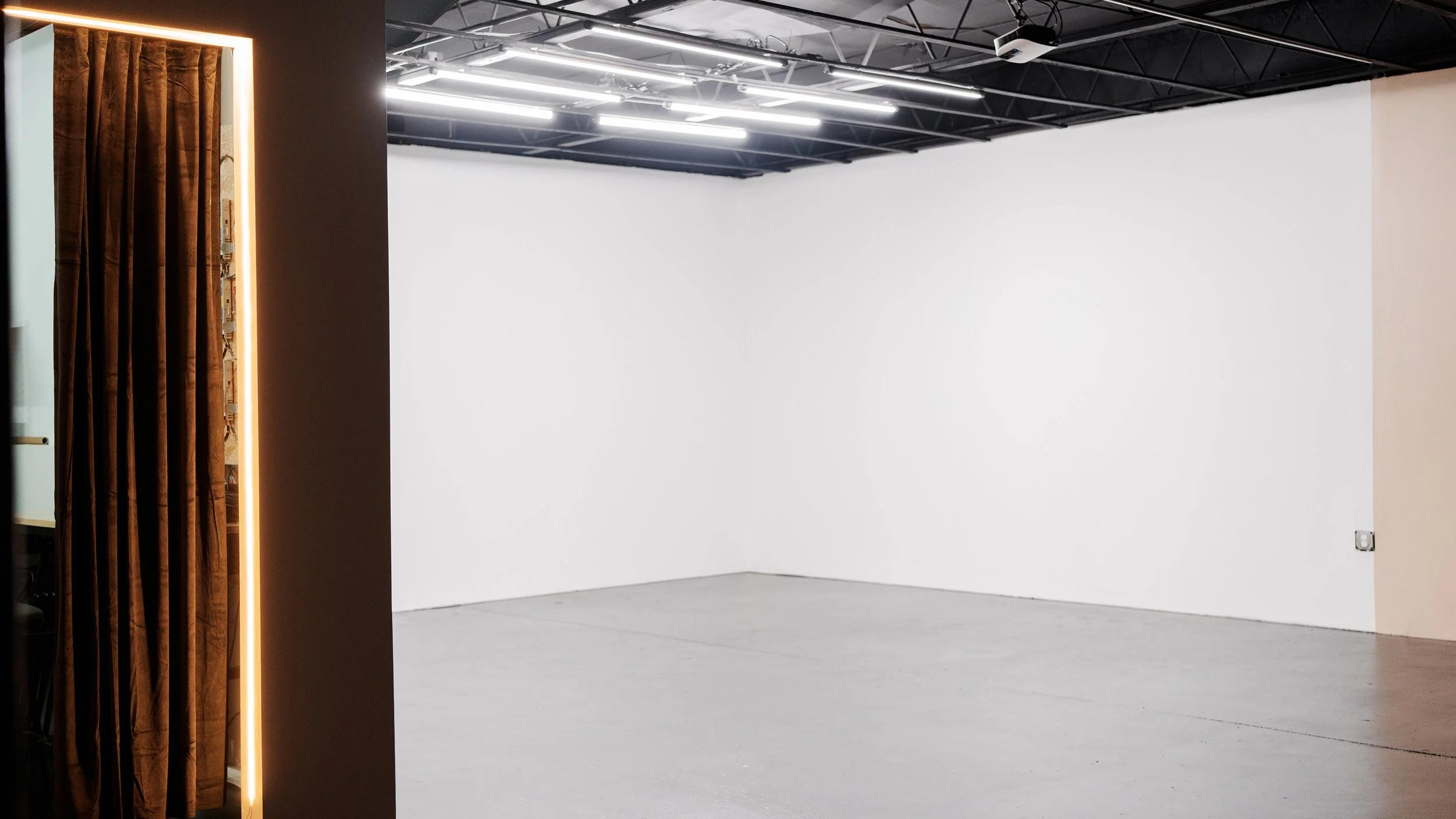 Empty gallery or exhibition space with white walls and a gray floor, partially visible brown curtains with a lighted frame on the left, and ceiling lights.