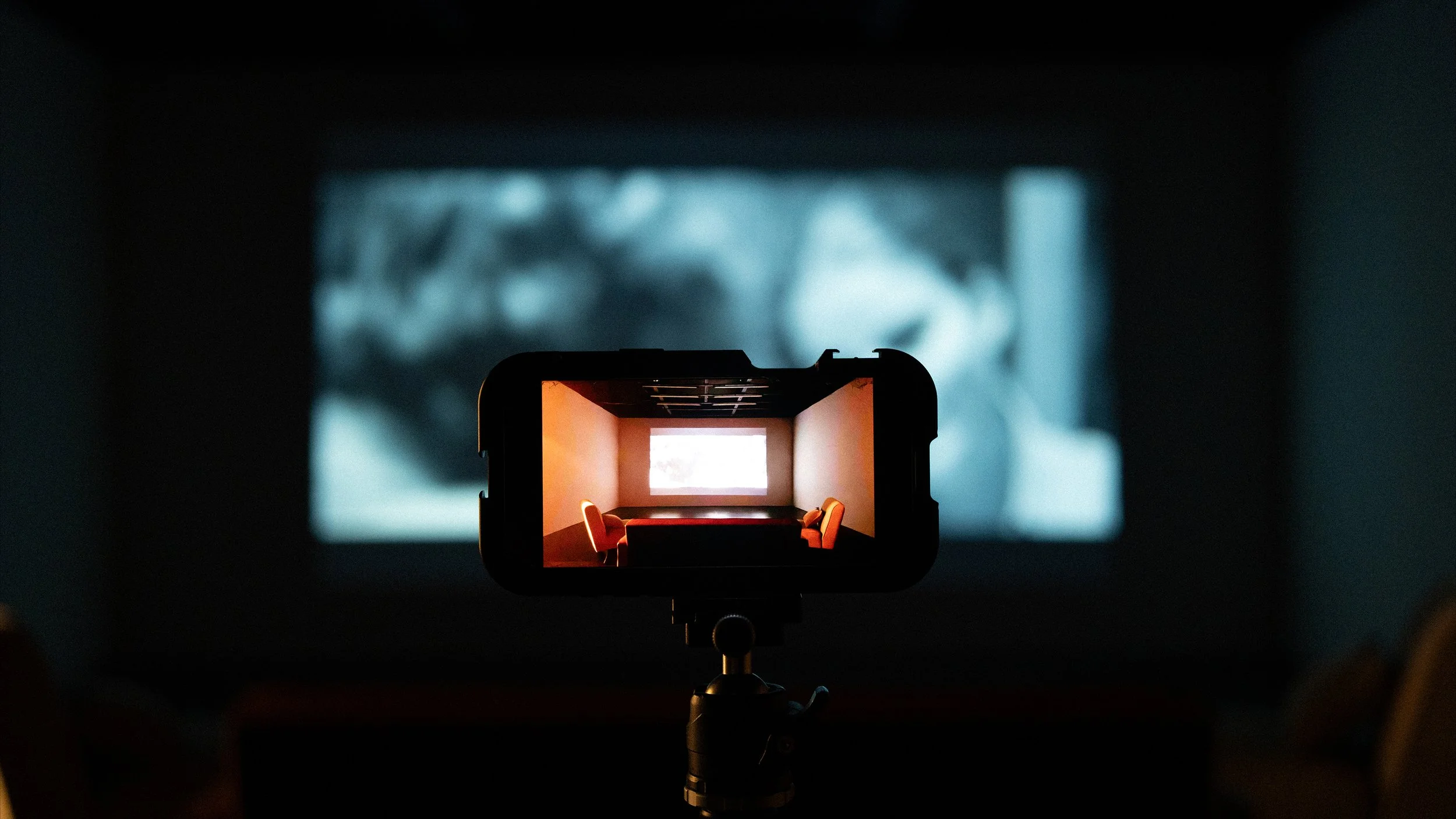 A camera capturing a room with a large screen on the wall and orange chairs arranged around a table, viewed through the camera's display.
