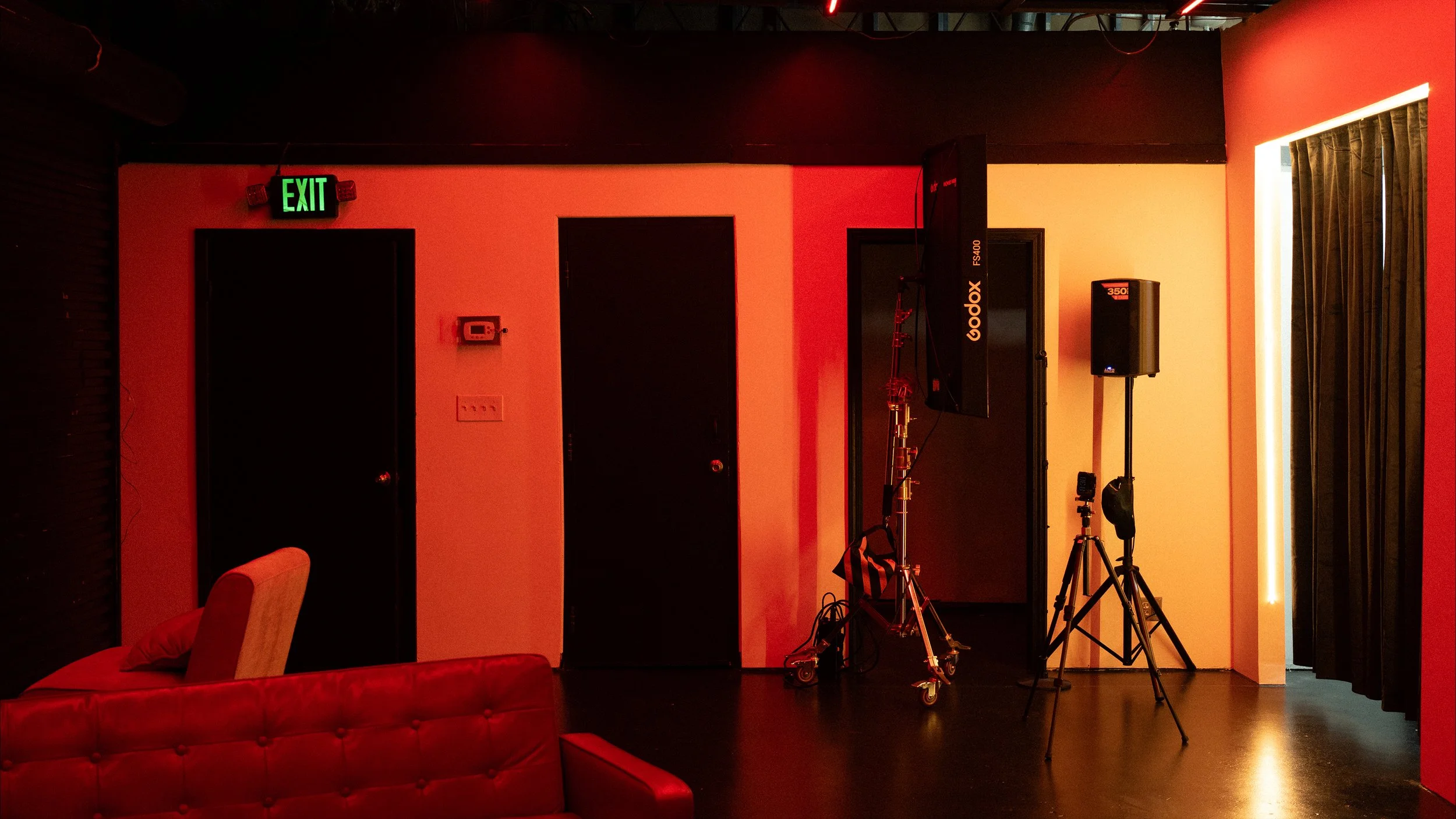 Indoor space with red leather sofa, black walls, mounted TV, professional lighting and camera equipment, and an exit sign.