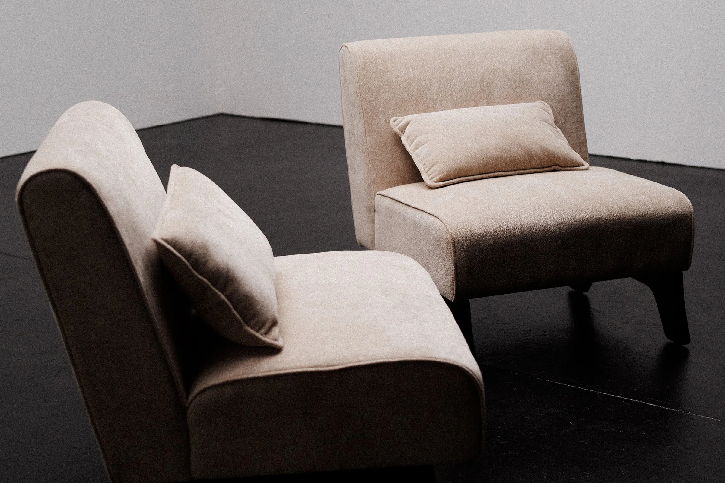 Two beige upholstered armchairs with cushions on a black floor against a plain light gray wall.