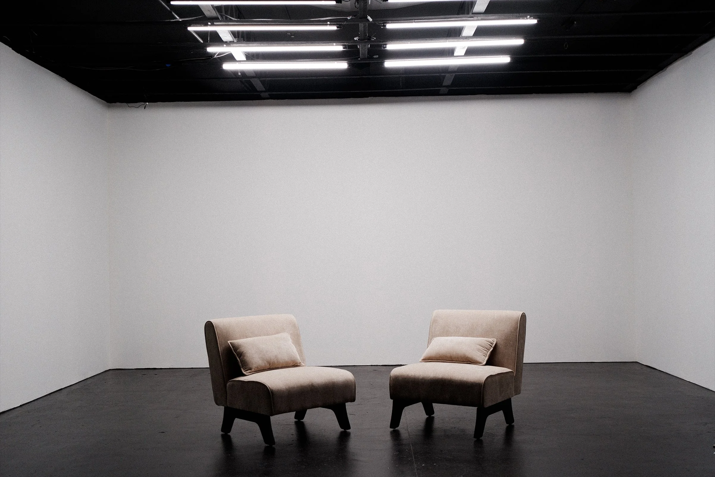 An empty studio or room with white walls and black ceiling, containing two beige upholstered armchairs with cushions, facing each other.