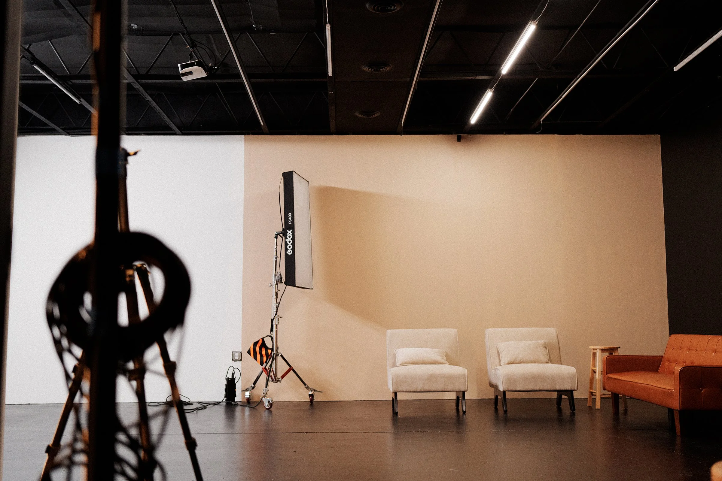 Photography studio with a beige wall, two white chairs, and an orange armchair, with photography equipment and lighting stands visible.