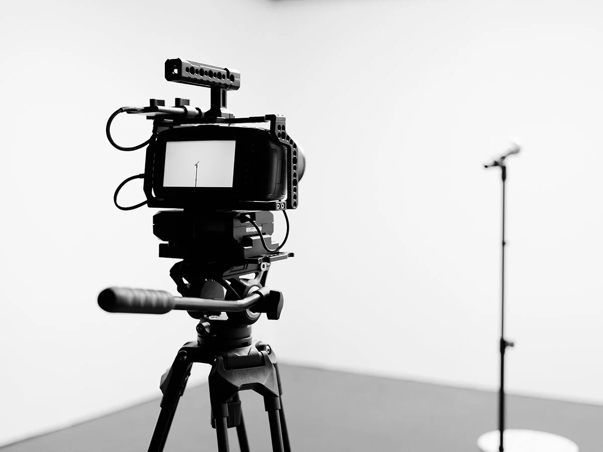A professional video camera set on a tripod, pointed towards a white background with a microphone stand in the distance.