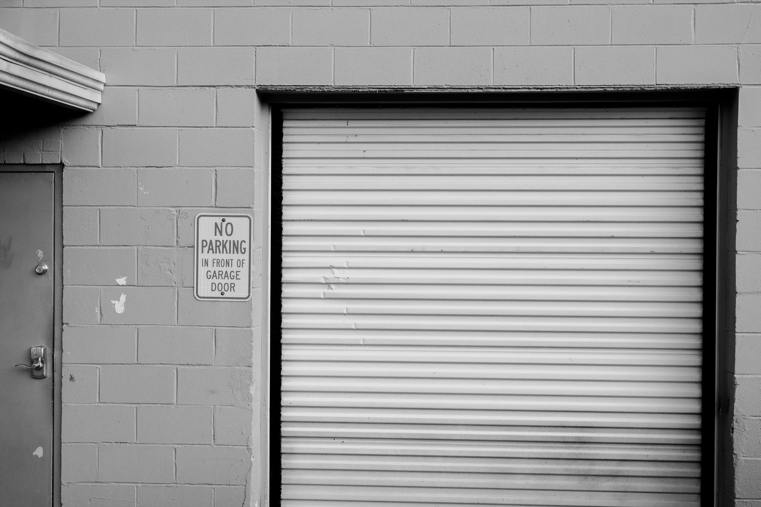 A black and white photo showing a garage door, a brick wall, and a no parking sign that reads "No Parking in Front of Garage Door."
