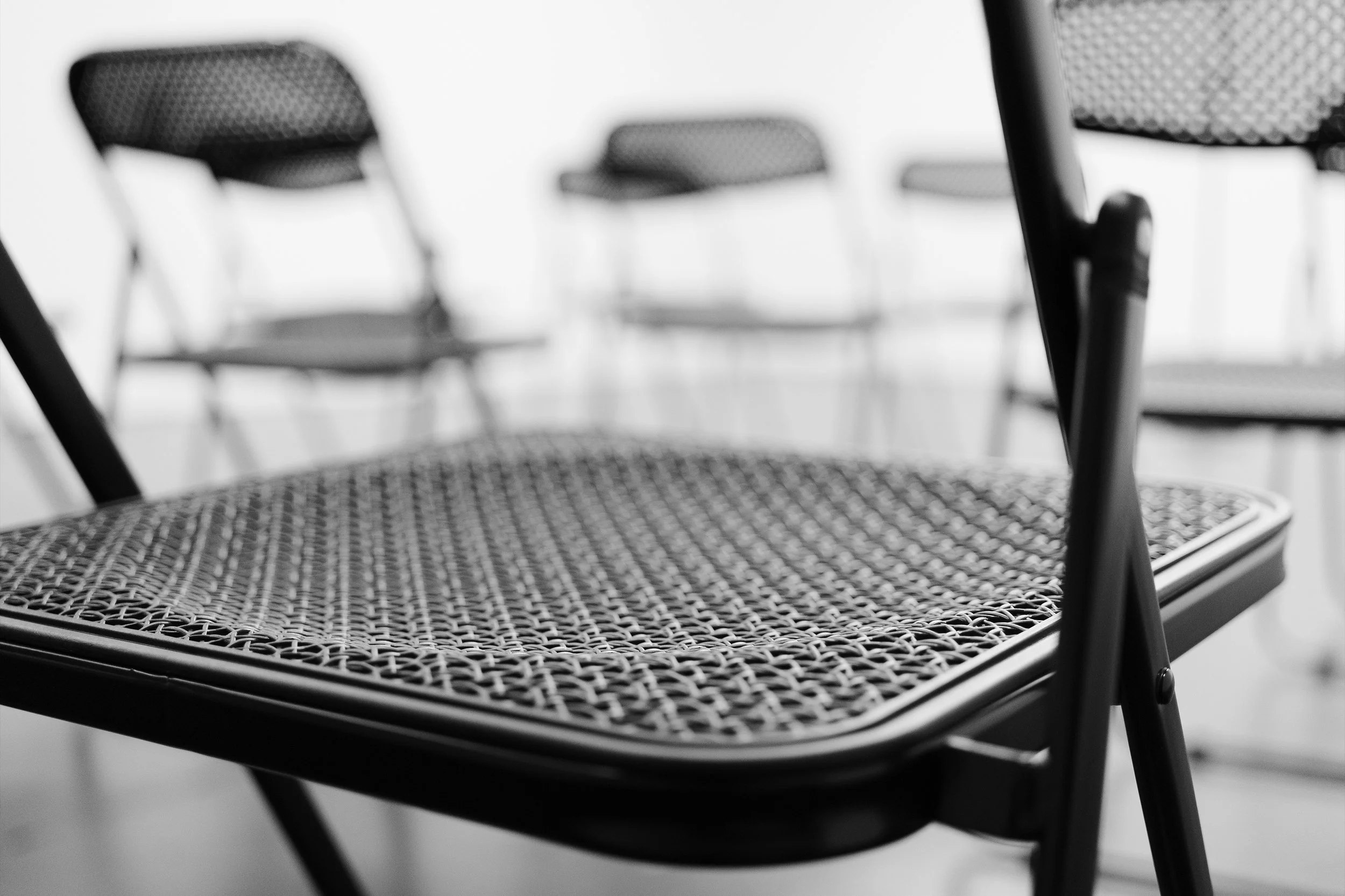 Close-up of a black metal chair with a woven mesh seat and backrest, with other similar chairs blurred in the background.