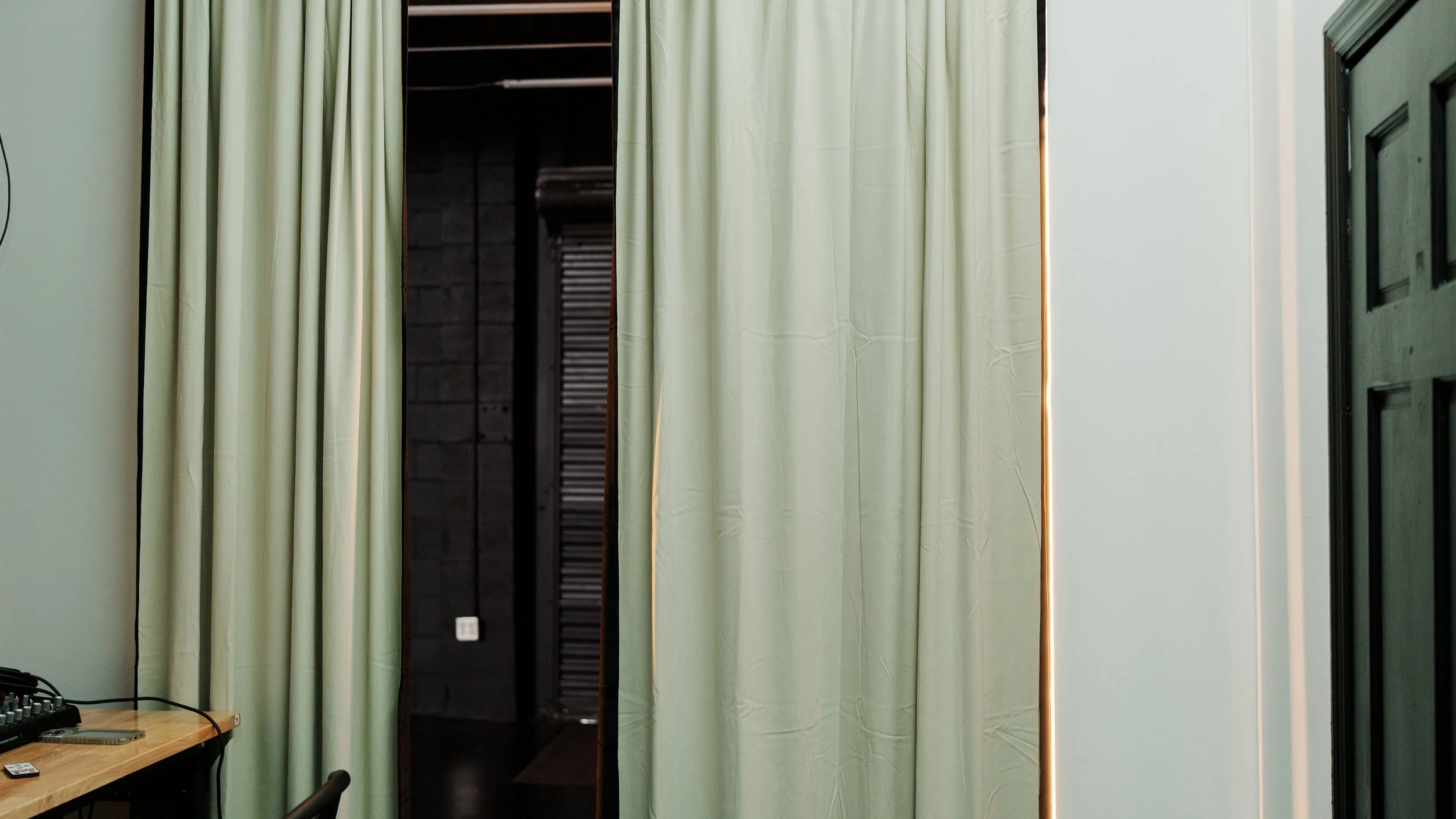 A room with cream-colored curtains hanging in front of a dark open space as a doorway, with a small portion of a wooden table with electronic devices on it visible on the left side.