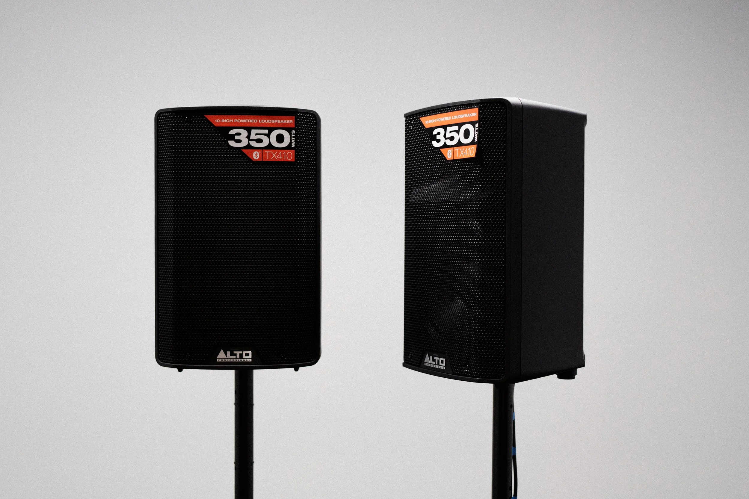 Two black Alto professional speakers with red and white labels indicating they are 350 watts and 10-inch powered loudspeakers, positioned against a plain light gray background.