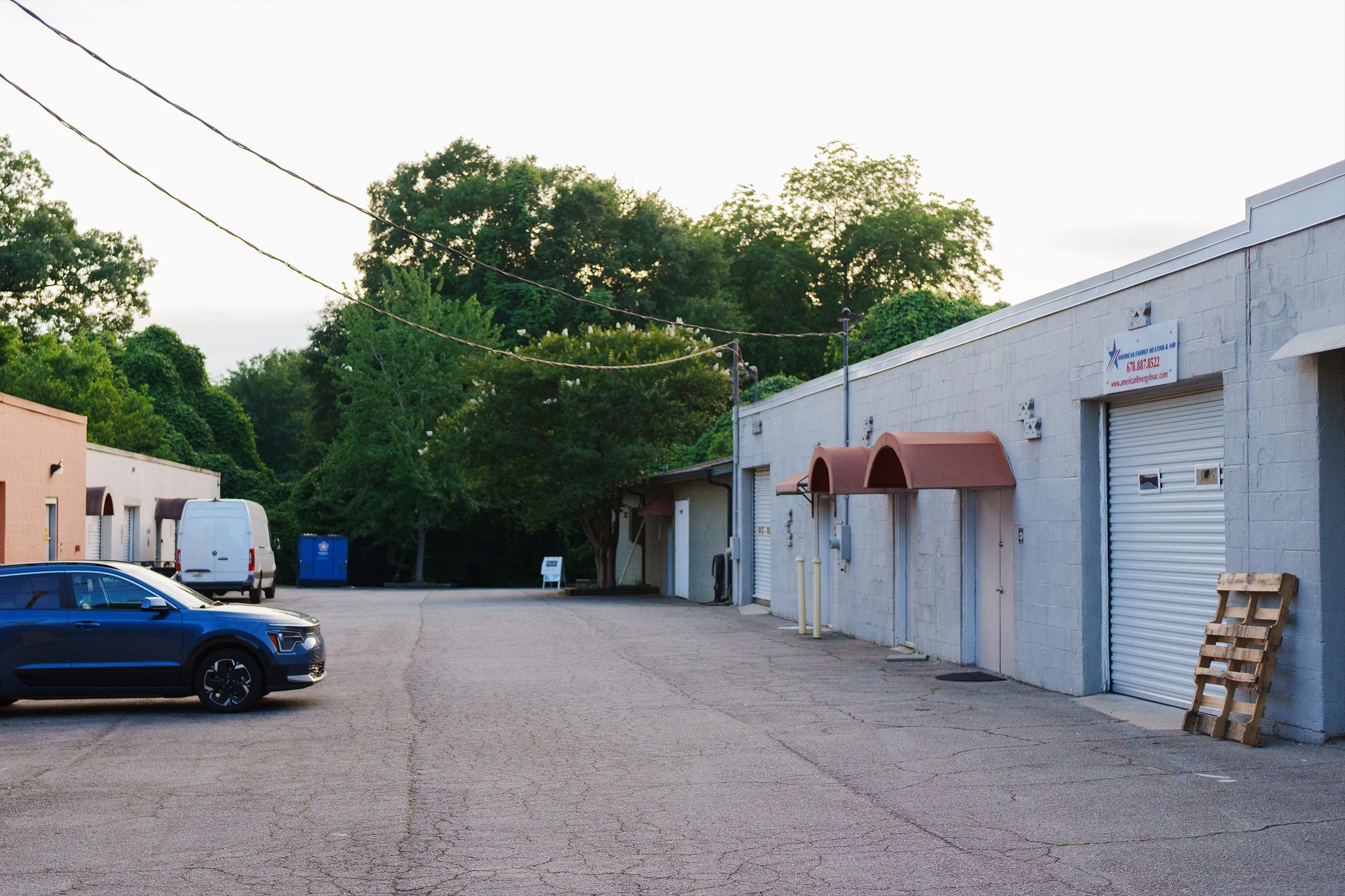 An outdoor parking lot with a few parked vehicles, including a blue SUV, a white van, and a dark-colored car. On the right side, there is a white industrial building with closed roll-up doors, small awnings, and a sign that reads 'American Energy Hea