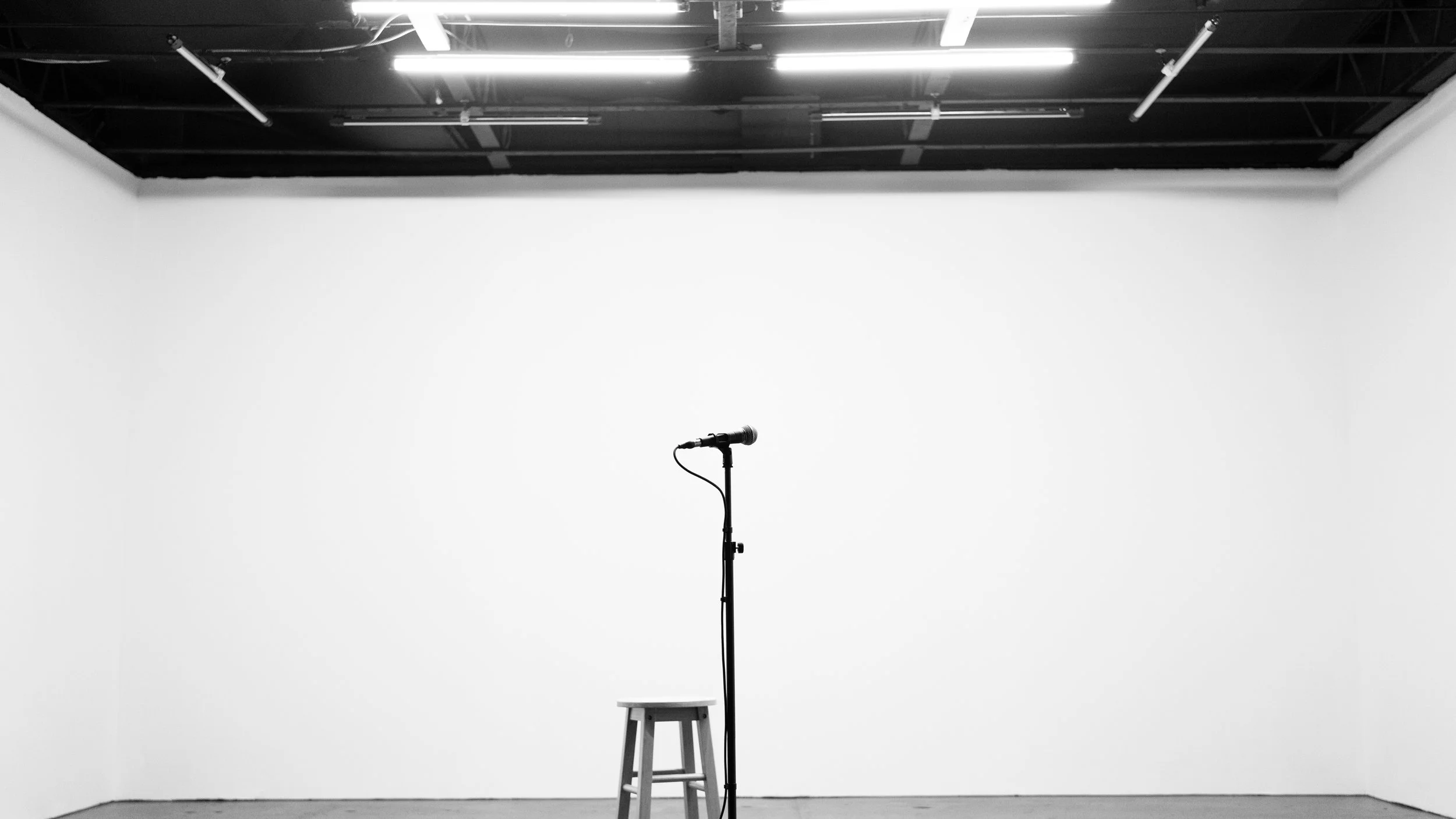 A black microphone on a stand, a wooden stool, and a stool are set up in an empty white-walled room with a black ceiling, ready for a performance or speech.