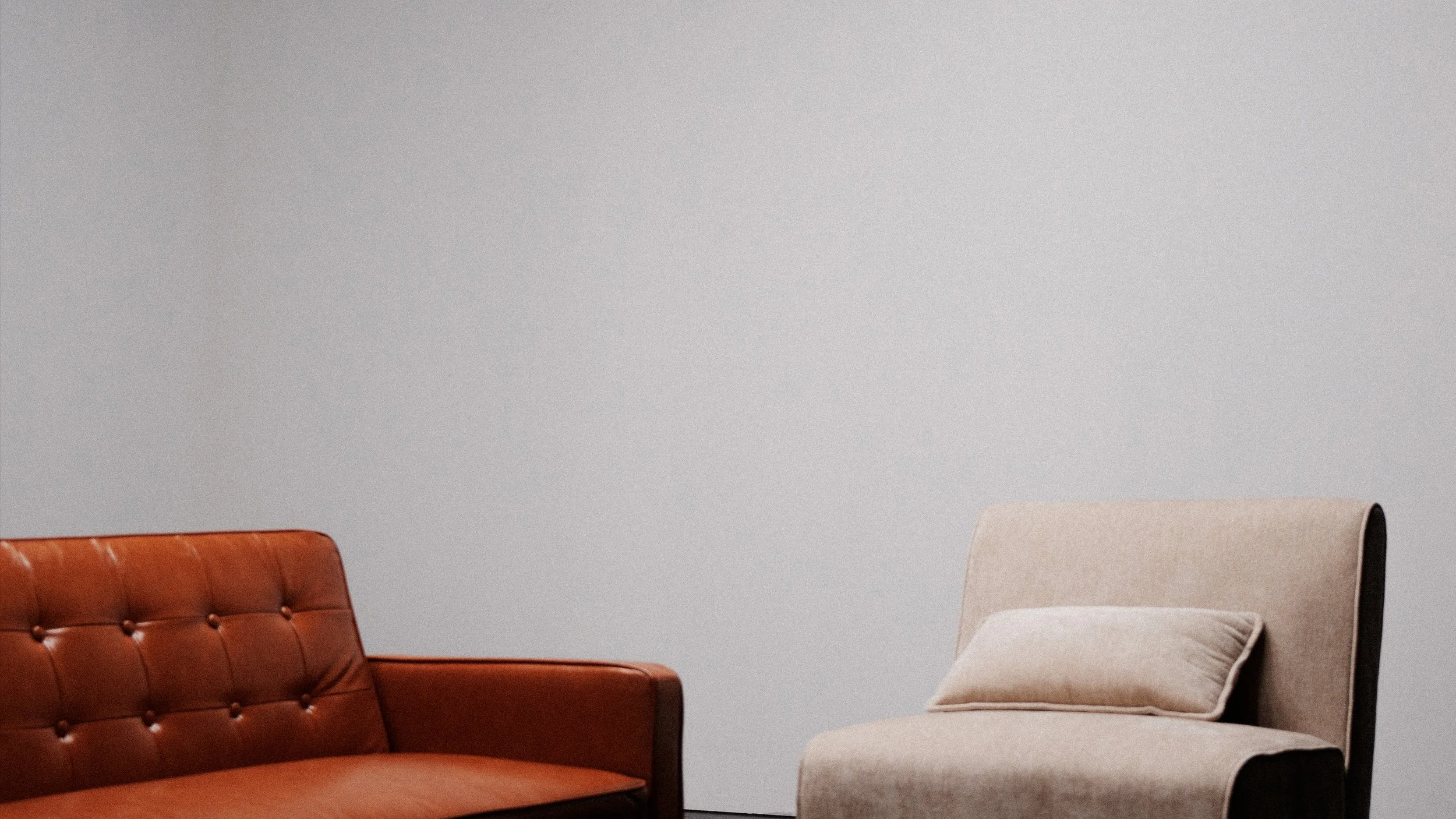 Two chairs, one brown leather and one beige fabric with a cushion, against a plain gray wall.