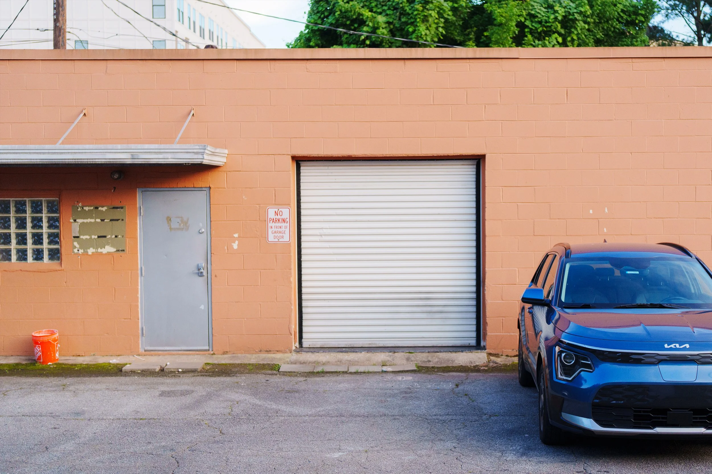 The image shows the front of a peach-colored building with a gray door, a large closed white roll-up garage door, and a small window with glass blocks. A red and white sign reads 'No Parking in Front of Garage Door.' An orange bucket is on the ground