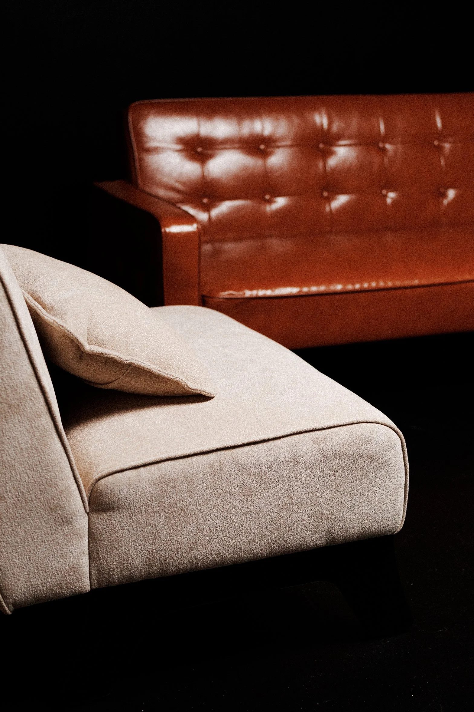 A beige upholstered couch with rounded armrests and a matching pillow, partially visible in the foreground, against a dark background. A brown leather tufted sofa is in the background.