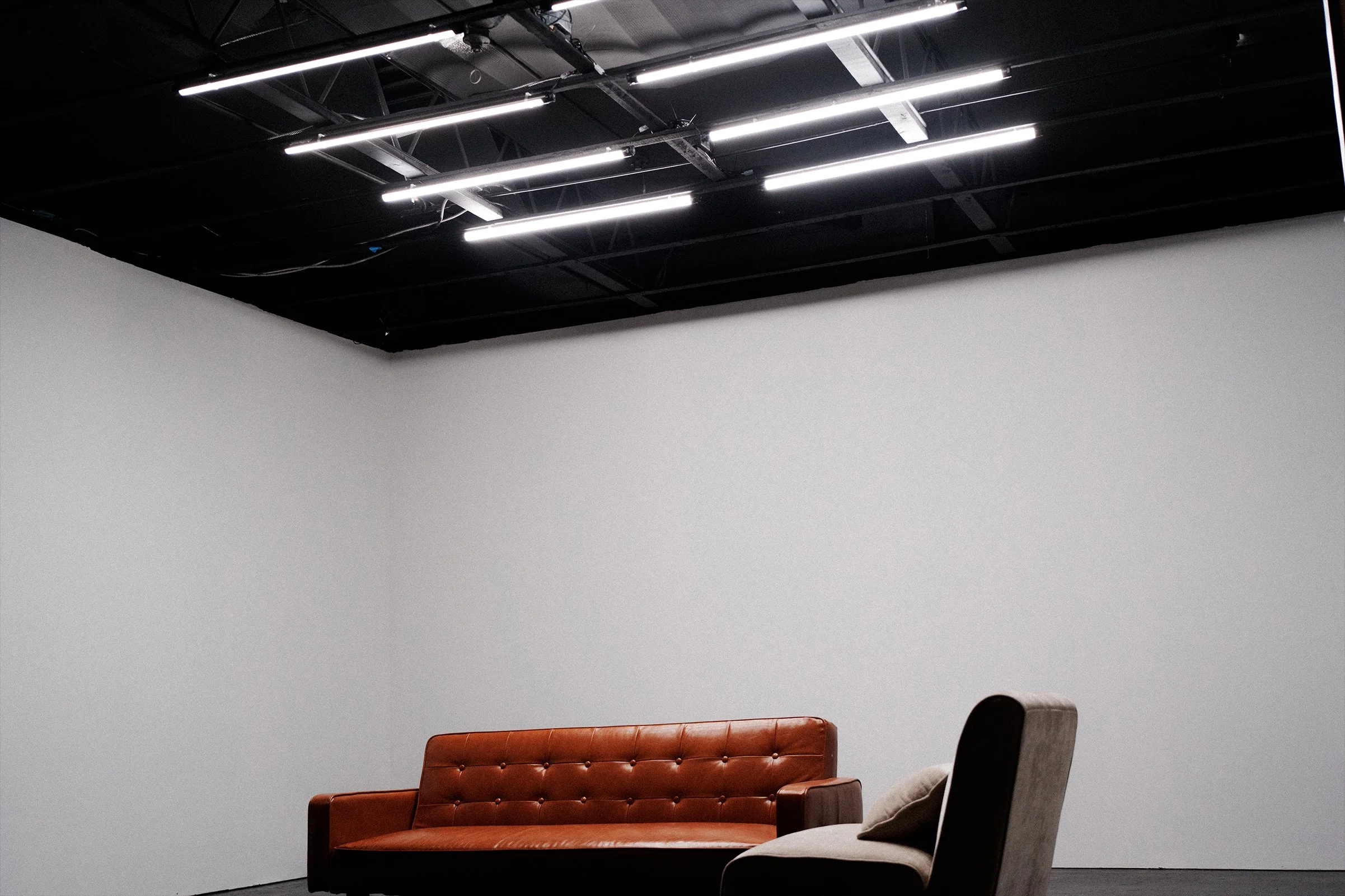 A brown leather sofa with a tufted backrest below a white wall, with a black chair and grey cushion nearby, in a room with a black ceiling and linear ceiling lights.