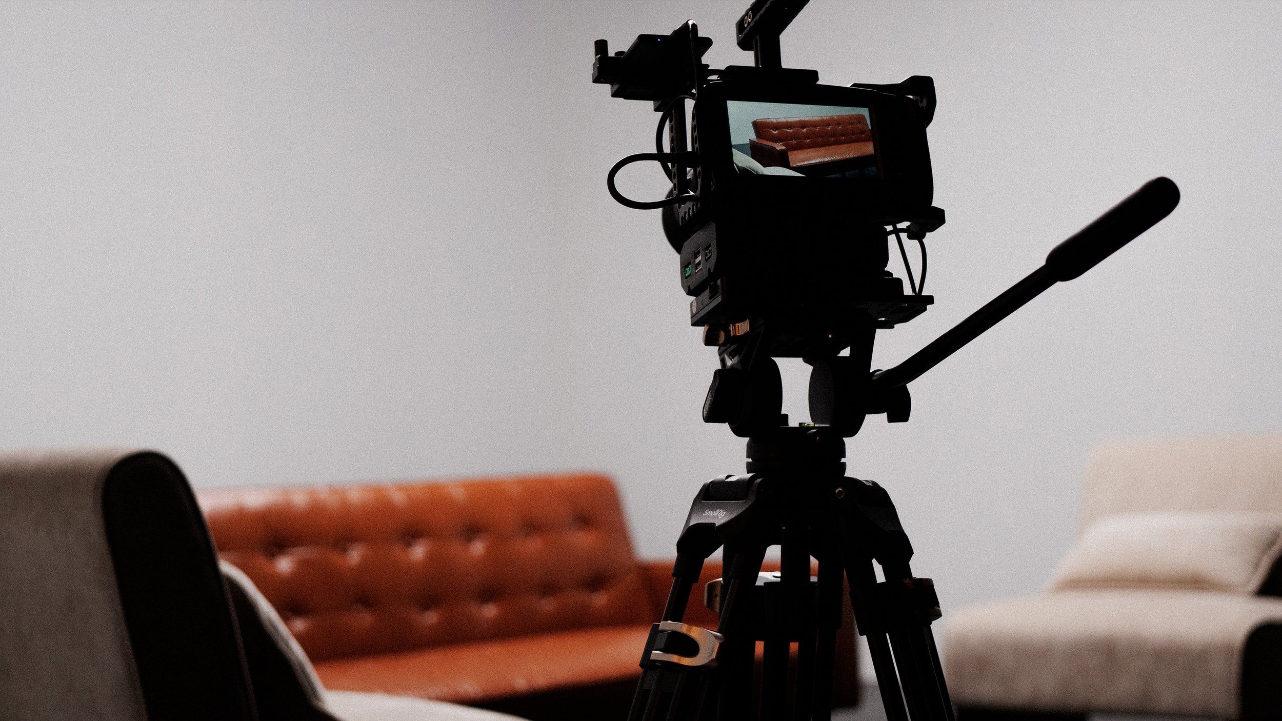A professional camera mounted on a tripod in a room with chairs, with a projection of a brown leather couch on the camera's screen.