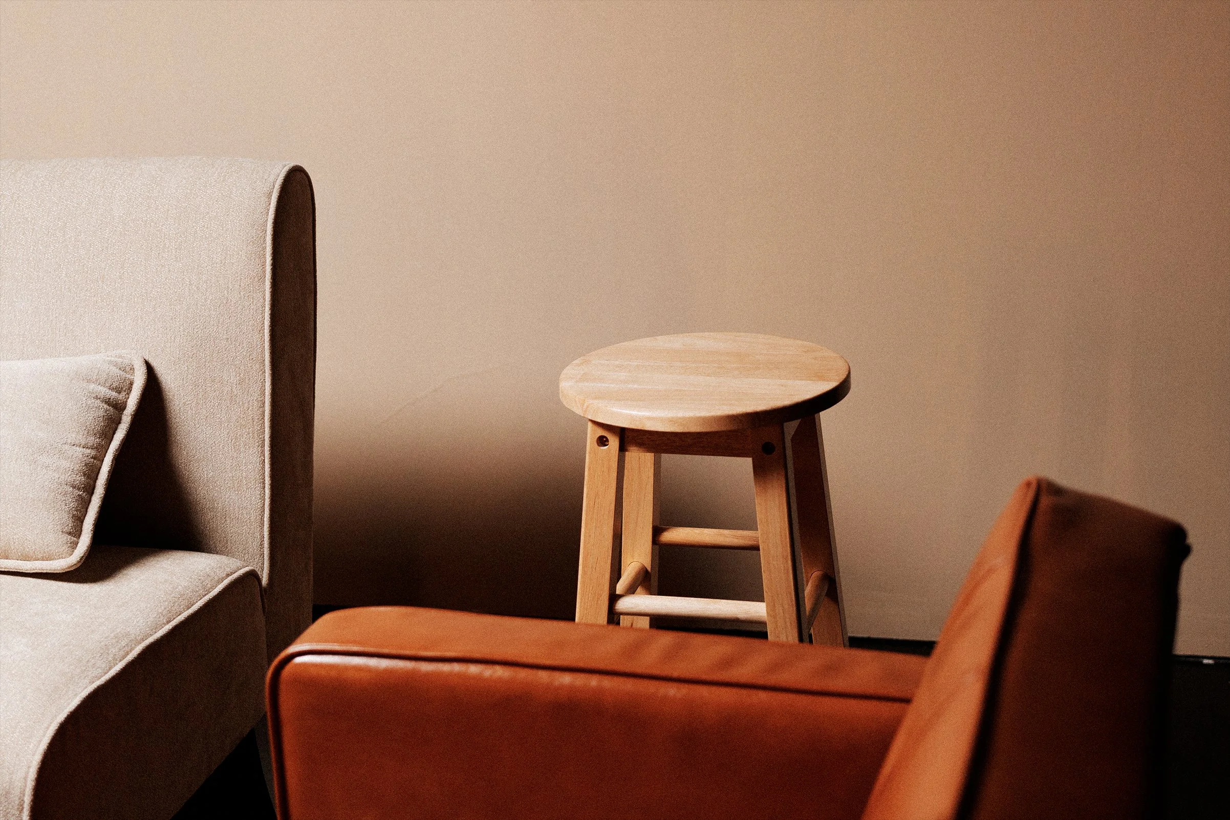 A beige upholstered armchair with a matching cushion, a round wooden stool, and a brown leather armchair against a plain beige wall.