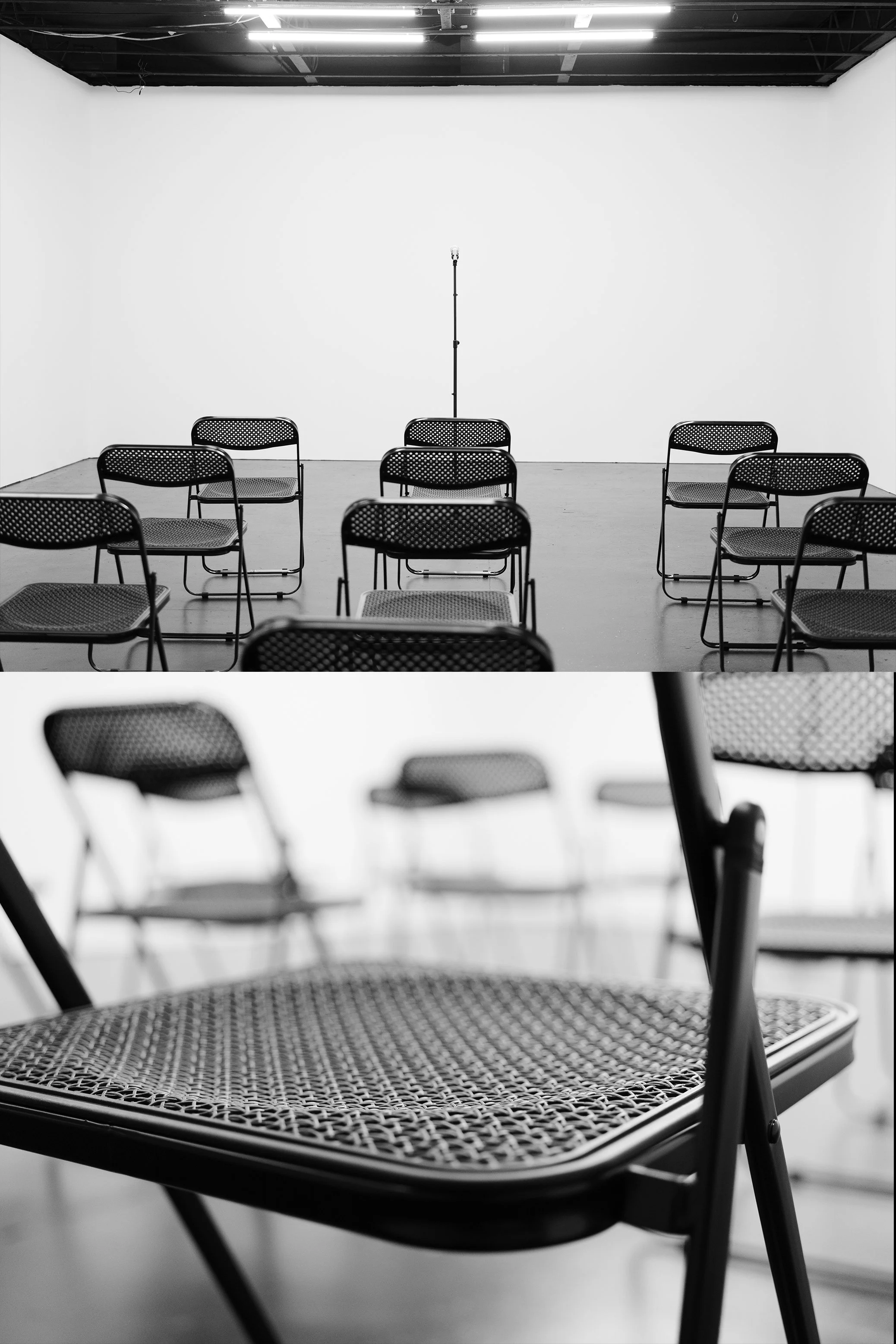 Empty black metal chairs with perforated seats and backs arranged in rows in a small, plain room with white walls and a ceiling with fluorescent lights, and a microphone stand at the front.
