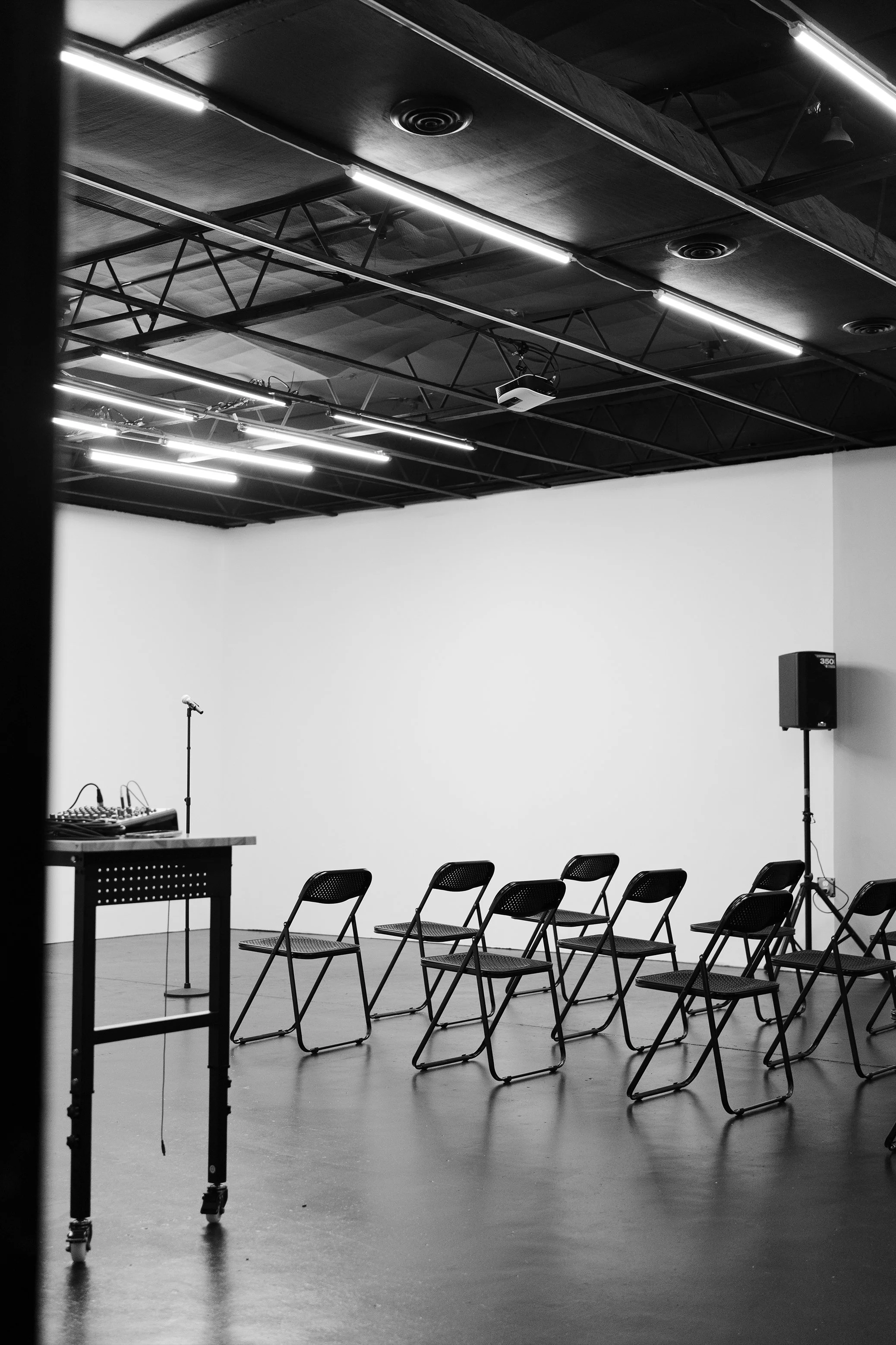 Empty stage with a microphone, chairs arranged in front, and audio equipment, set in a room with black ceiling, fluorescent lighting, and a white wall.