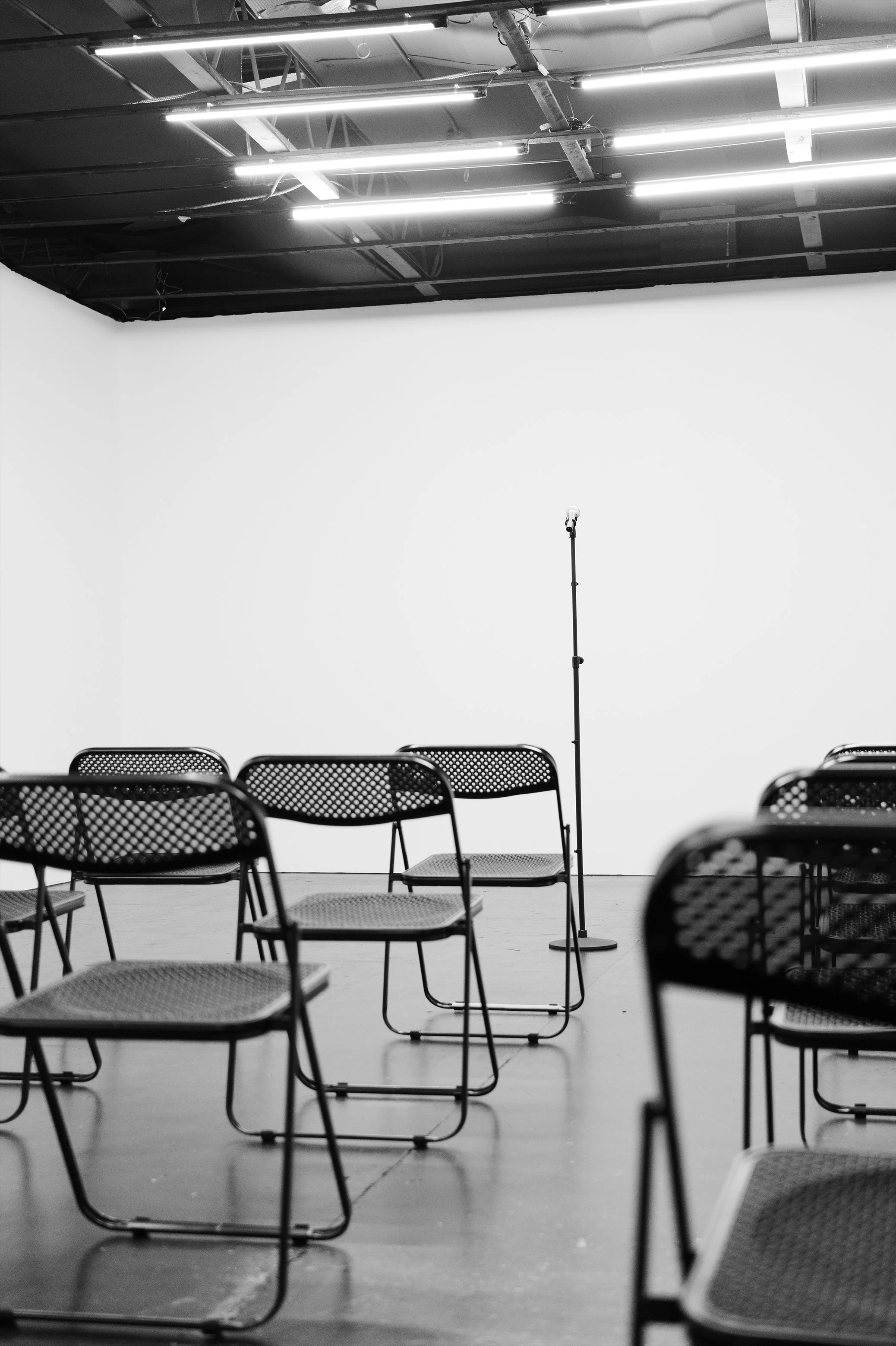 Empty chairs set up in rows facing a white wall, with a microphone stand and overhead fluorescent lights in a large, open room.