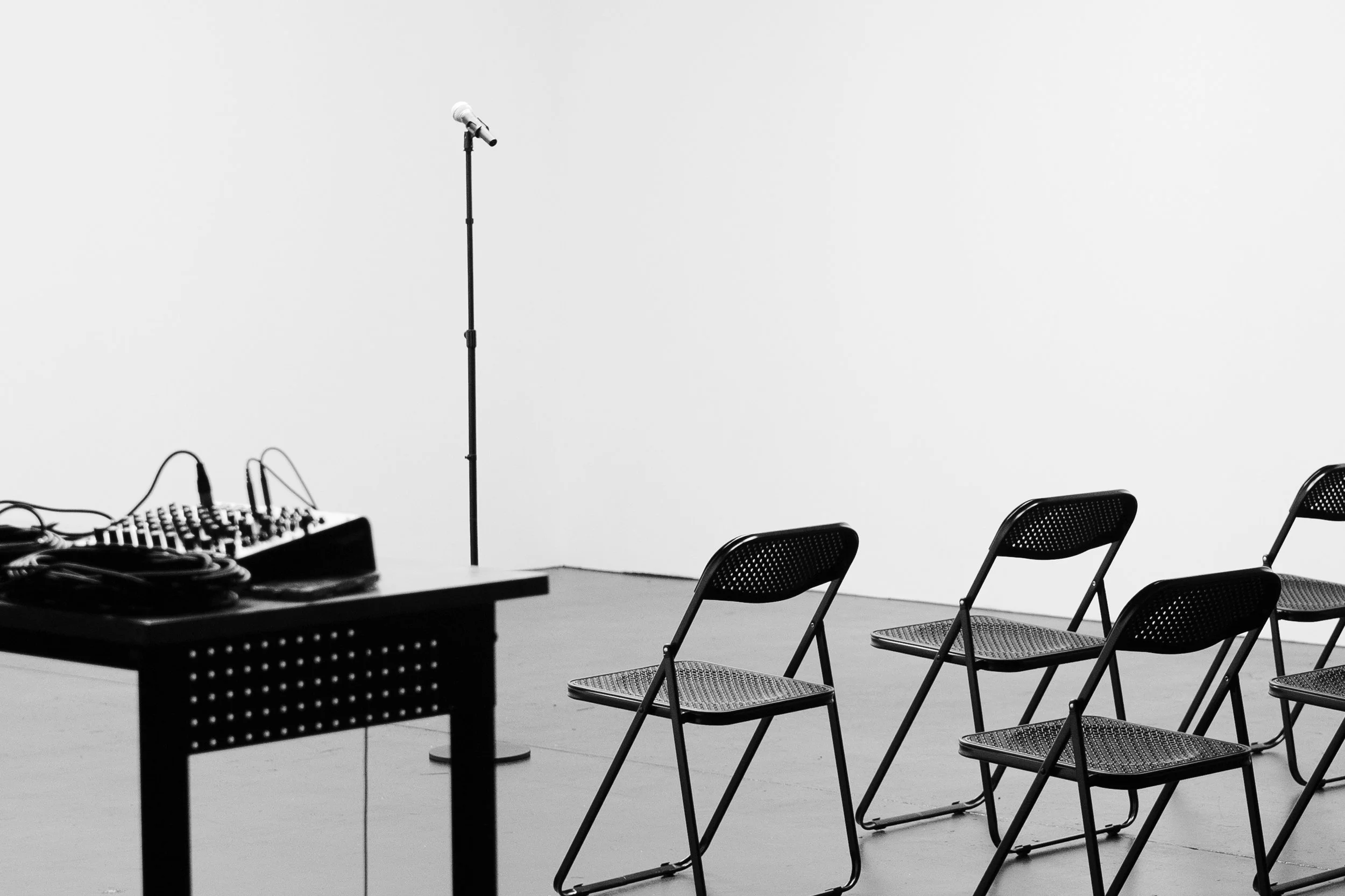 Empty chairs arranged in a row, near a table holding audio equipment, in front of a plain white wall, with a microphone on a stand positioned in front of the chairs.