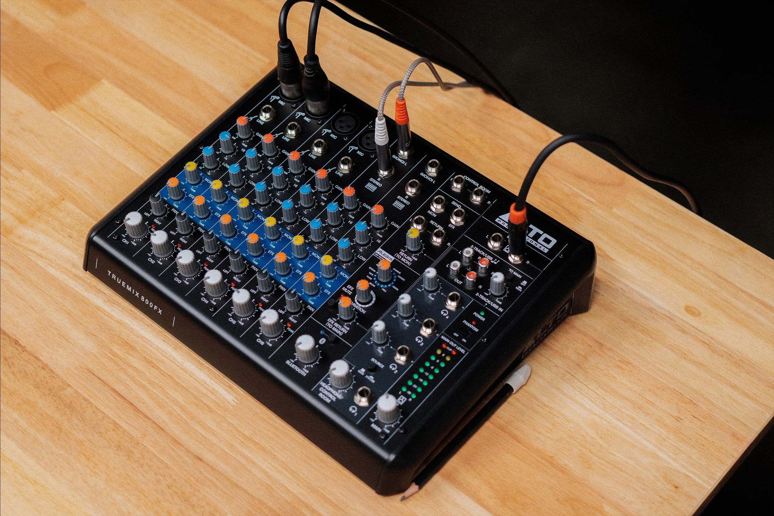 An audio mixing console with various knobs, sliders, and input/output ports on a wooden surface, connected to multiple cables.