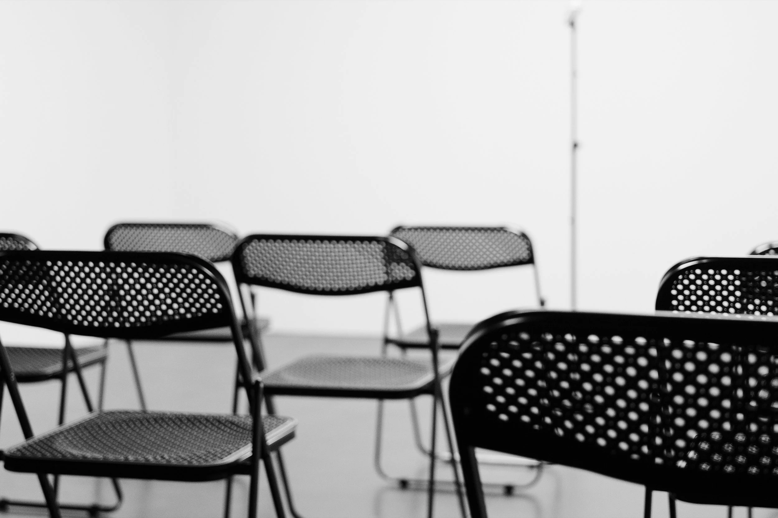 Multiple black metal mesh chairs arranged in rows against a plain white wall, with a tall standing lamp in the background.