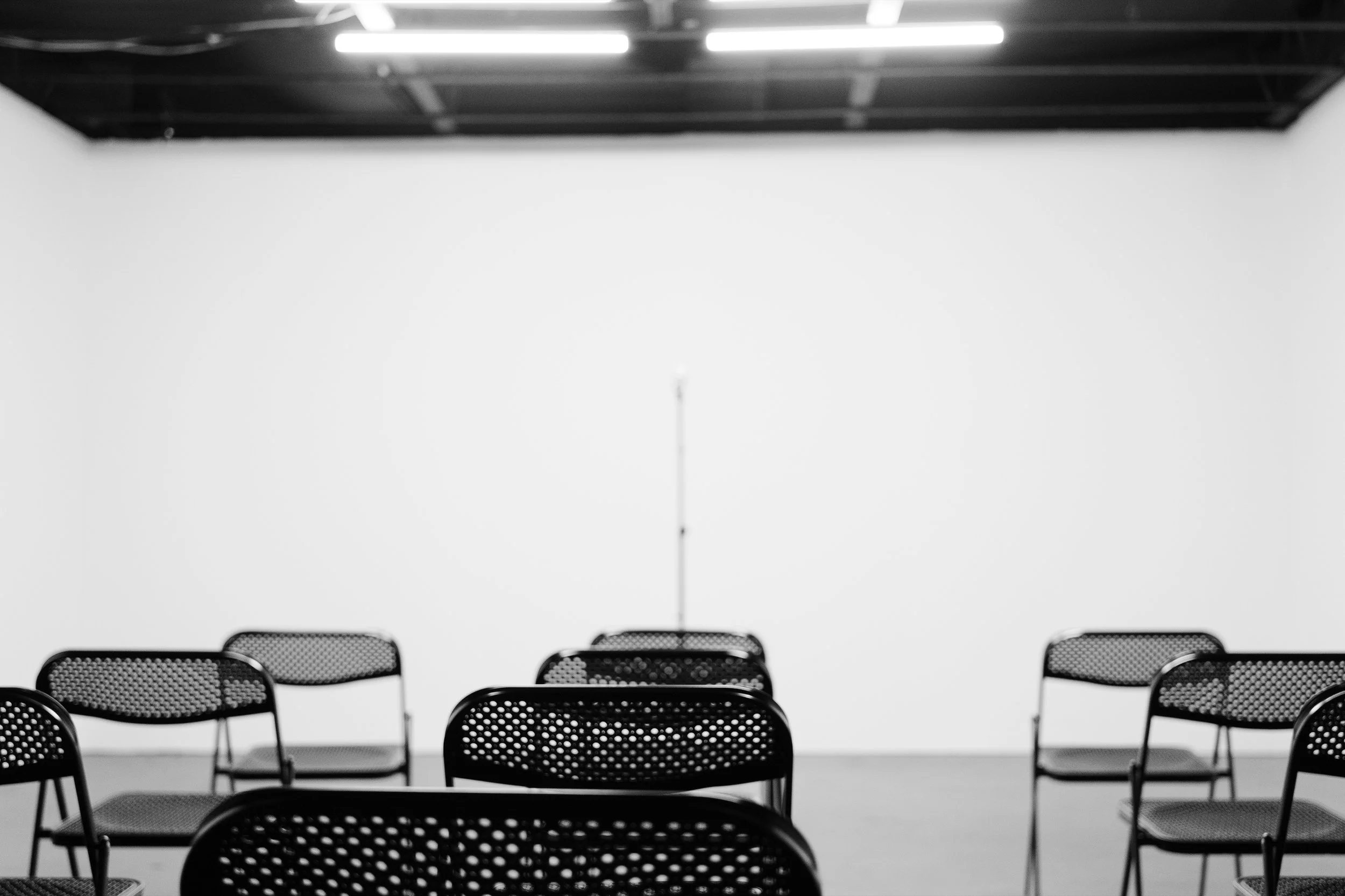 Empty black mesh chairs arranged in rows facing a white wall in a room with a black ceiling and fluorescent lighting.