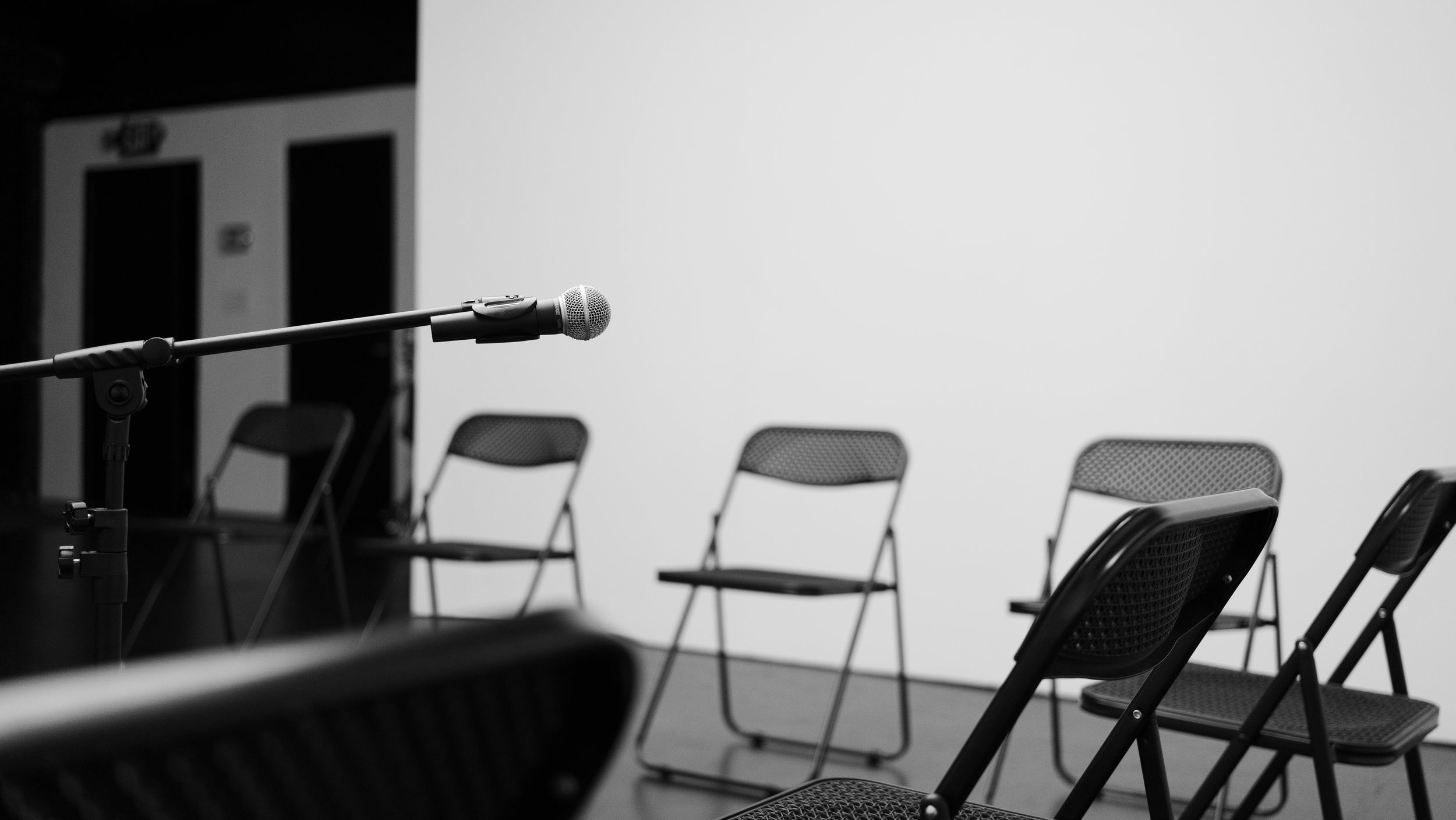 Empty chairs arranged in a row during a panel or presentation with a microphone on a stand in the foreground against a plain wall.