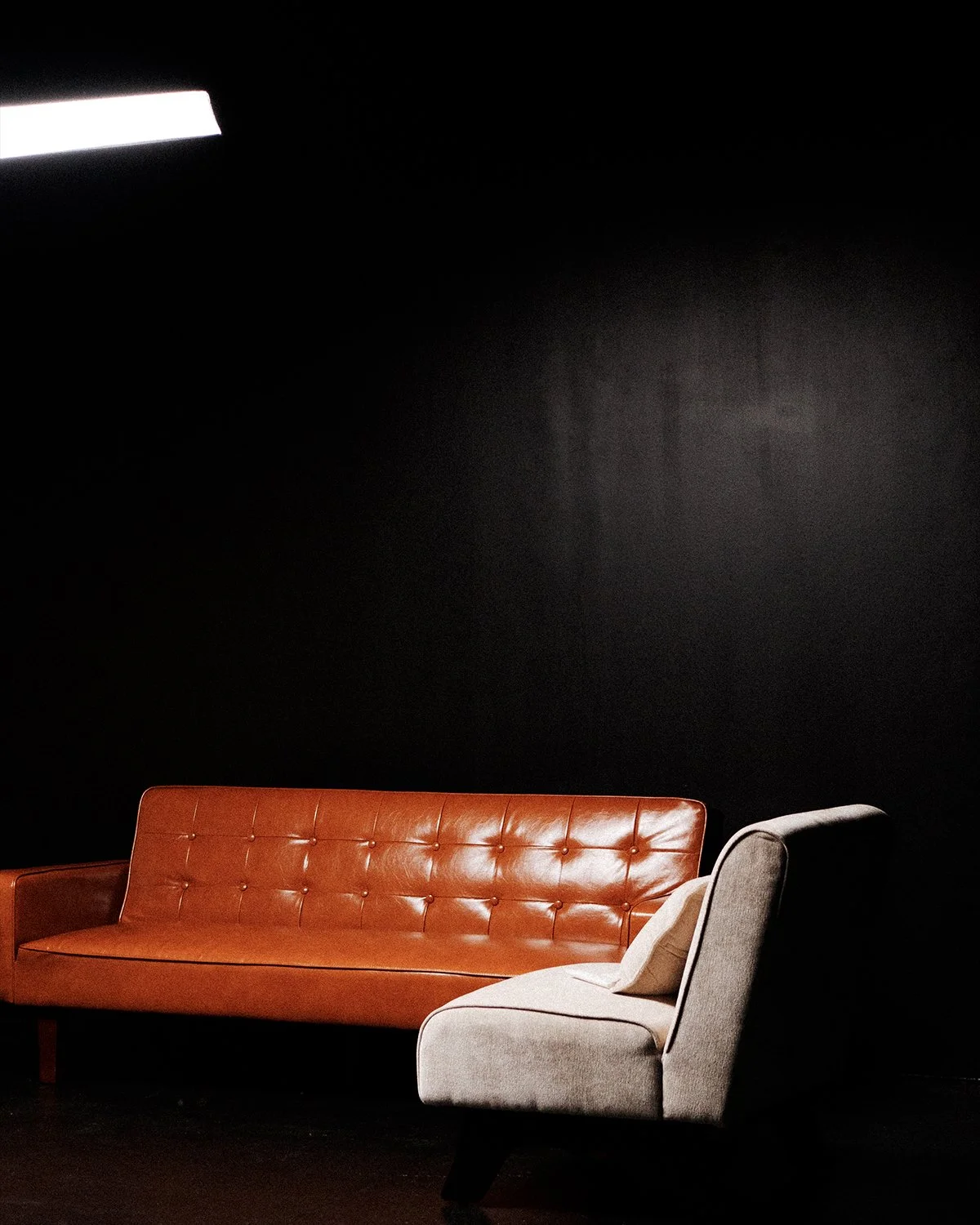 A minimalist interior scene with a burnt orange leather sofa and a light gray armchair against a black wall.